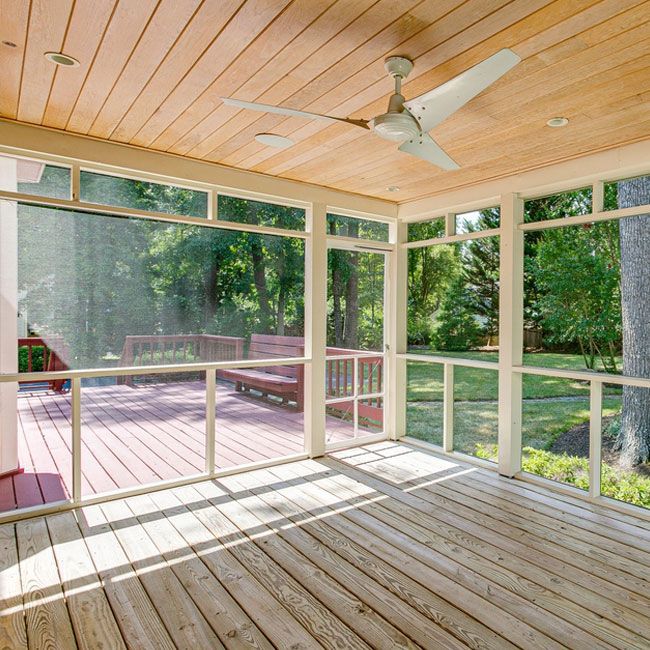 Sunlit screened porch with wooden floor, ceiling fan, and view of a deck and green yard