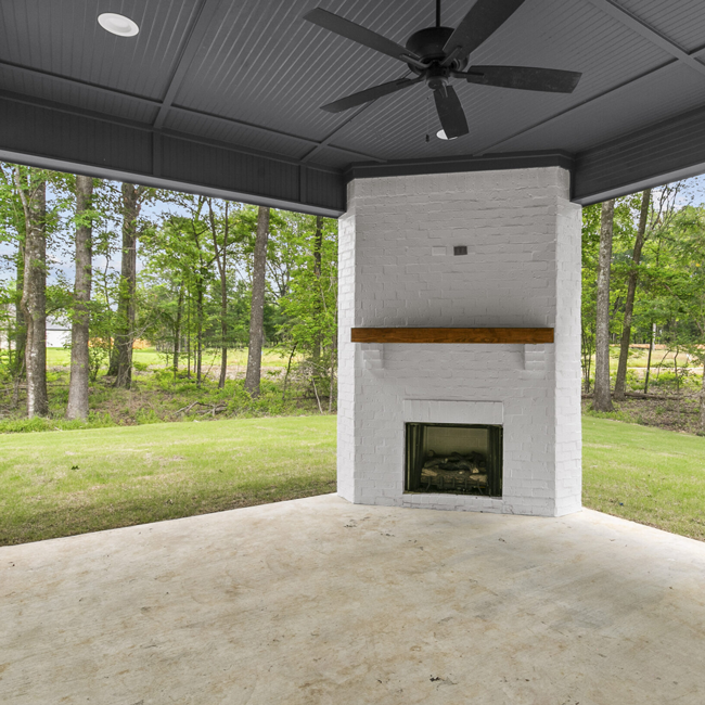 Covered patio with a white fireplace, concrete floor, ceiling fan, and wooded backyard view
