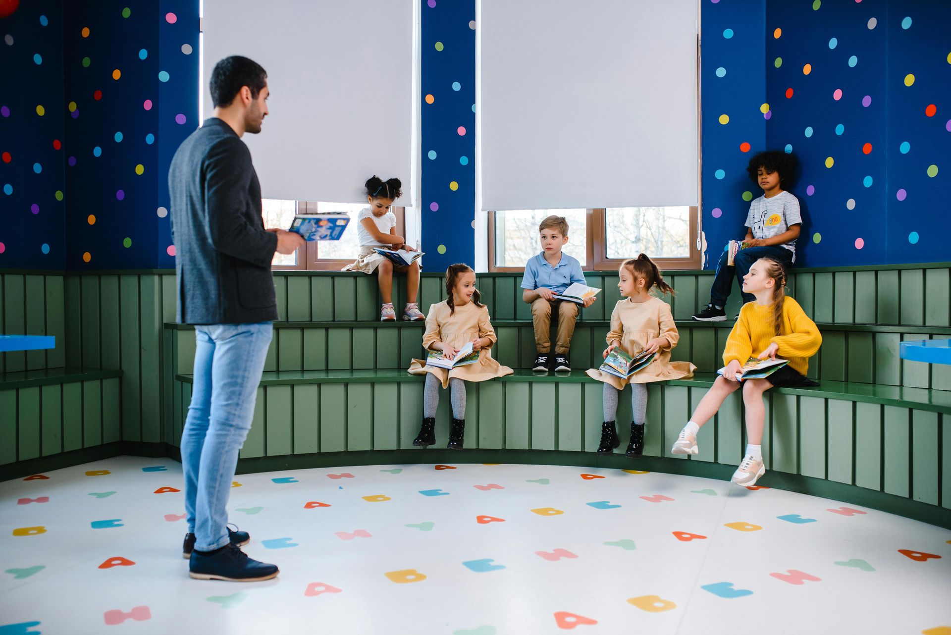 A man is standing in front of a group of children sitting on steps in a classroom.