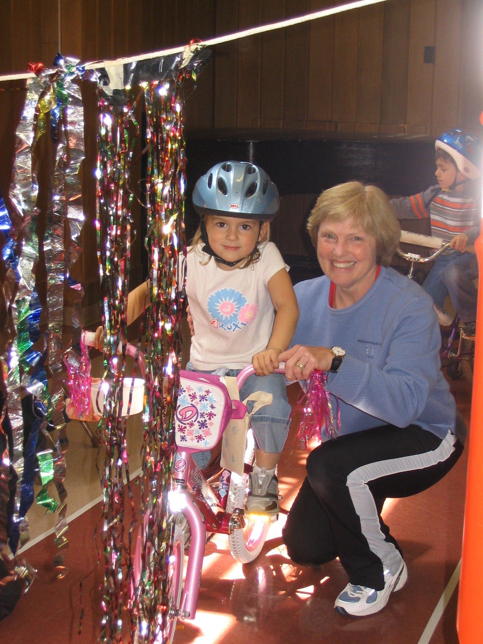 Two people smiling on a decorated indoor ride, one wearing a helmet, with colorful streamers and lights.