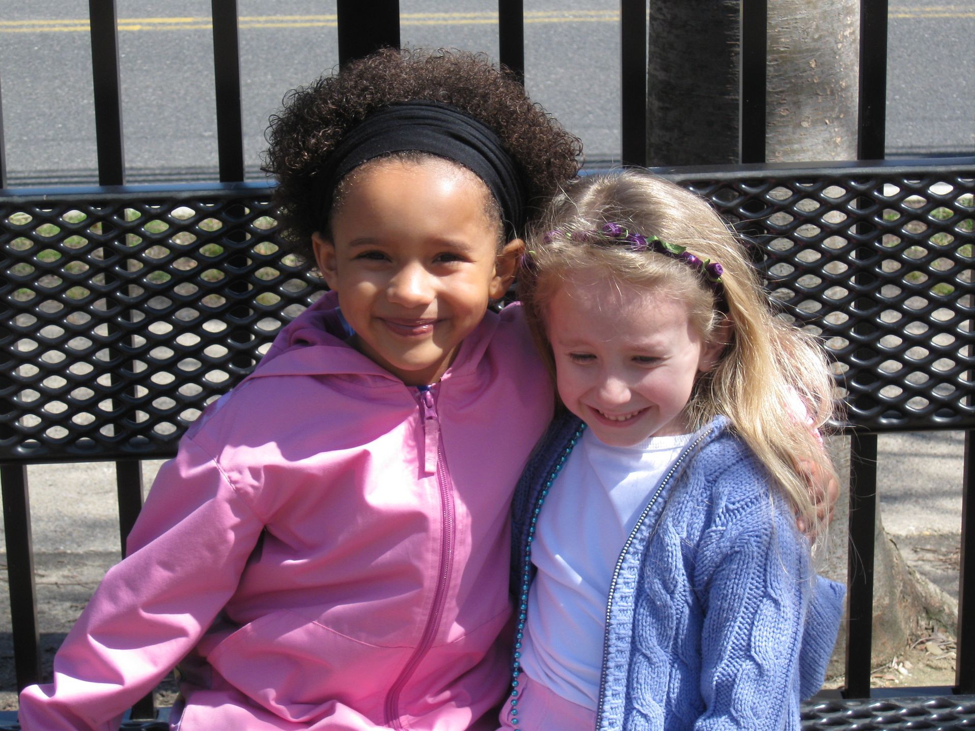 Two children smiling and hugging on a bench outdoors, one in a pink jacket and one in a blue sweater