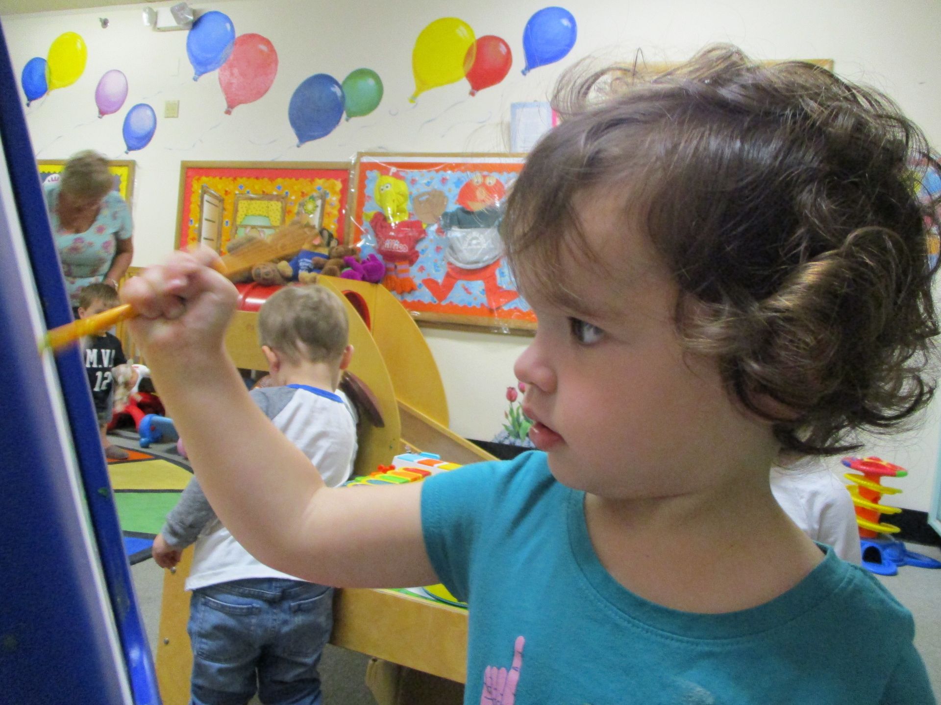 Child painting at an easel in a colorful classroom with balloons and other children in the background