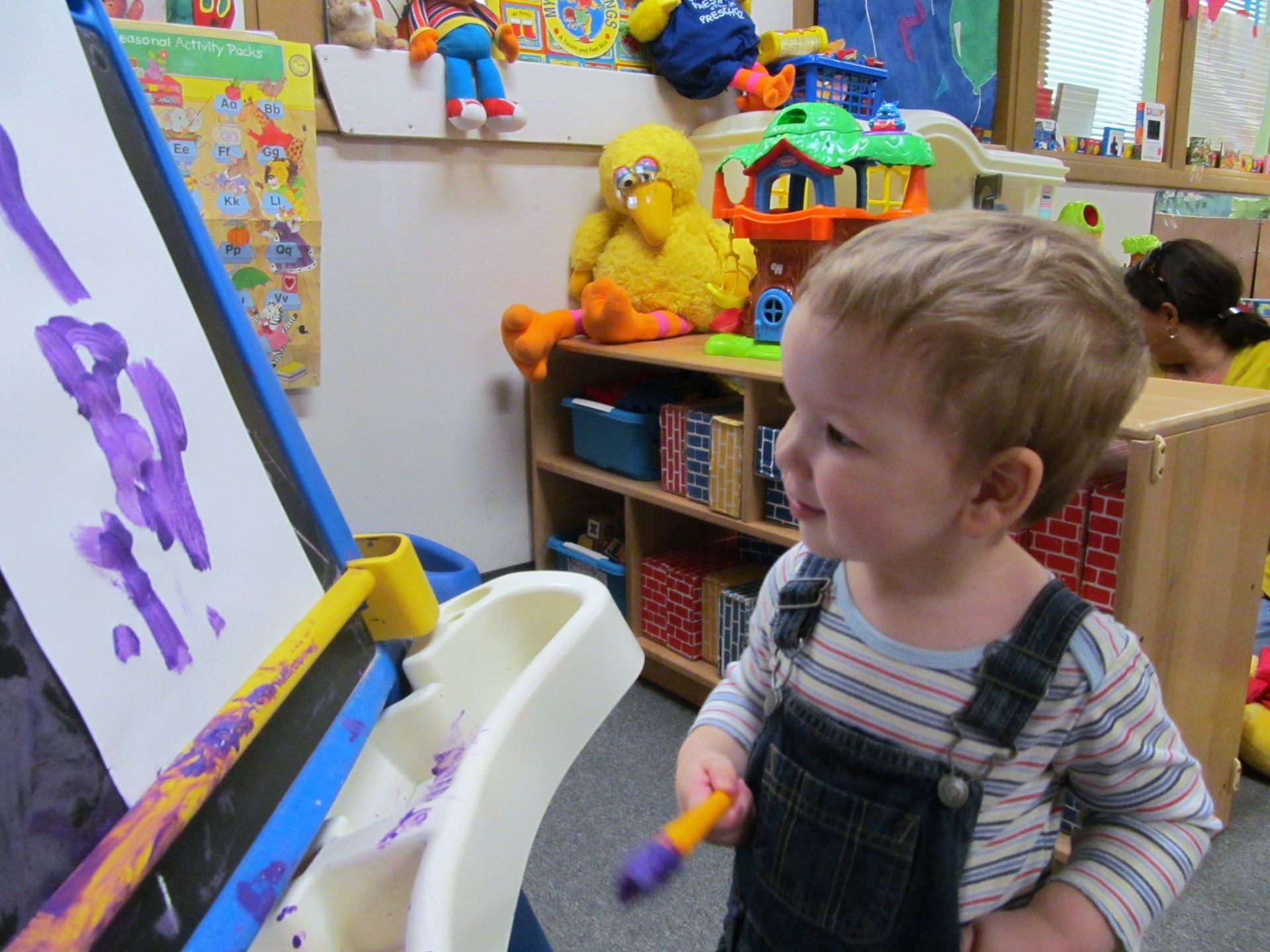 Child painting at an easel in a colorful classroom, holding a brush and smiling.