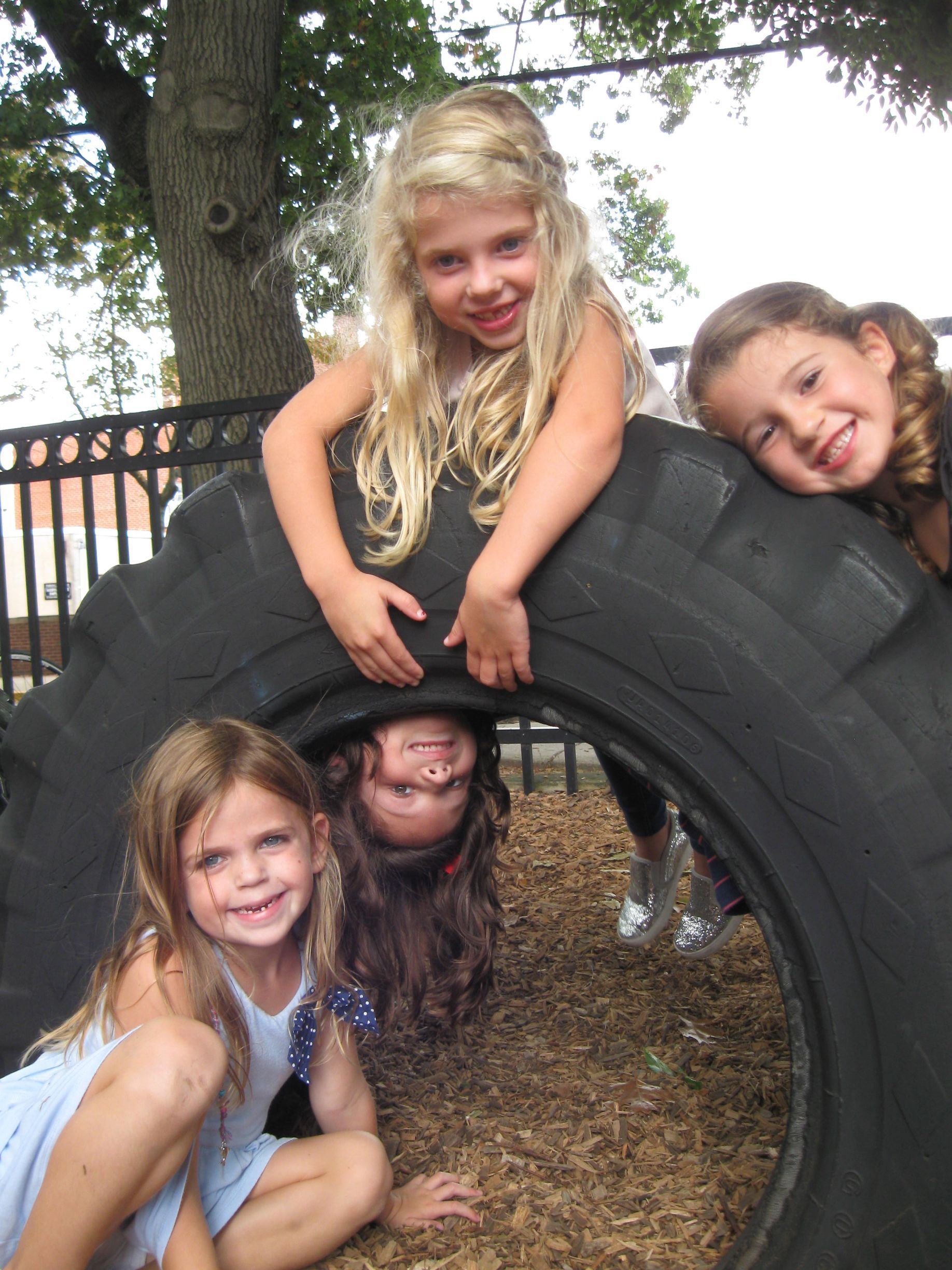 Four children smiling and hanging from a large tire outdoors near a tree