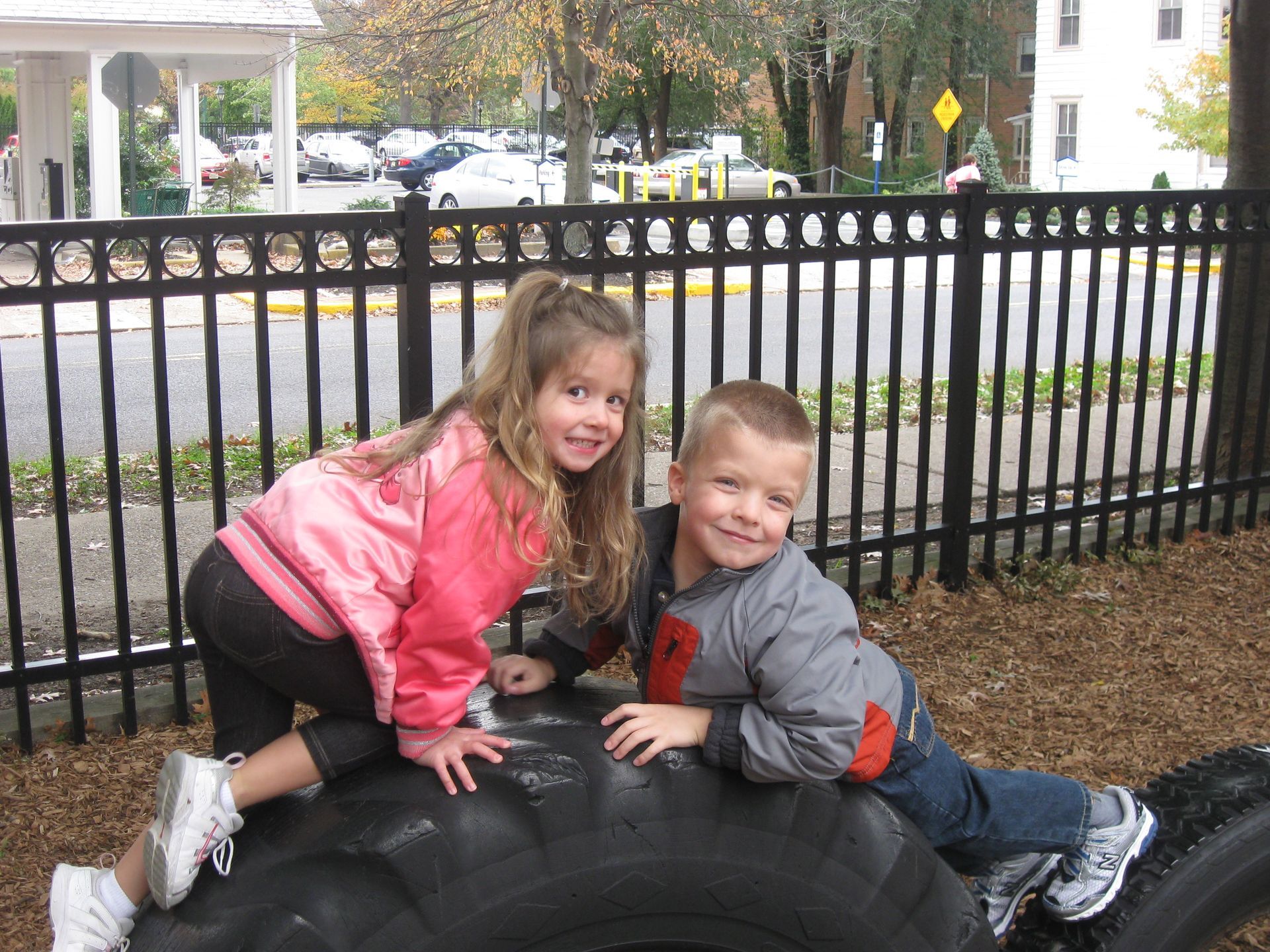 Two smiling children climbing on a large tire outdoors by a black fence