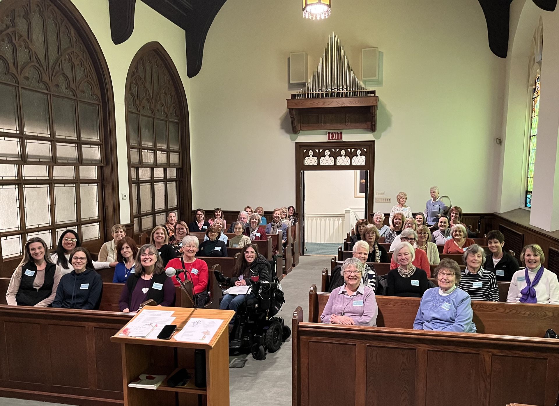 Audience seated in a church or lecture hall, facing a speaker’s podium at the front.
