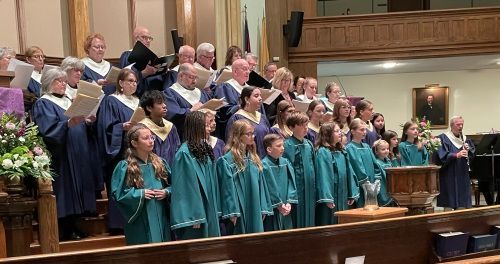 Choir members in teal robes stand on church steps during a performance, with seated singers behind them.