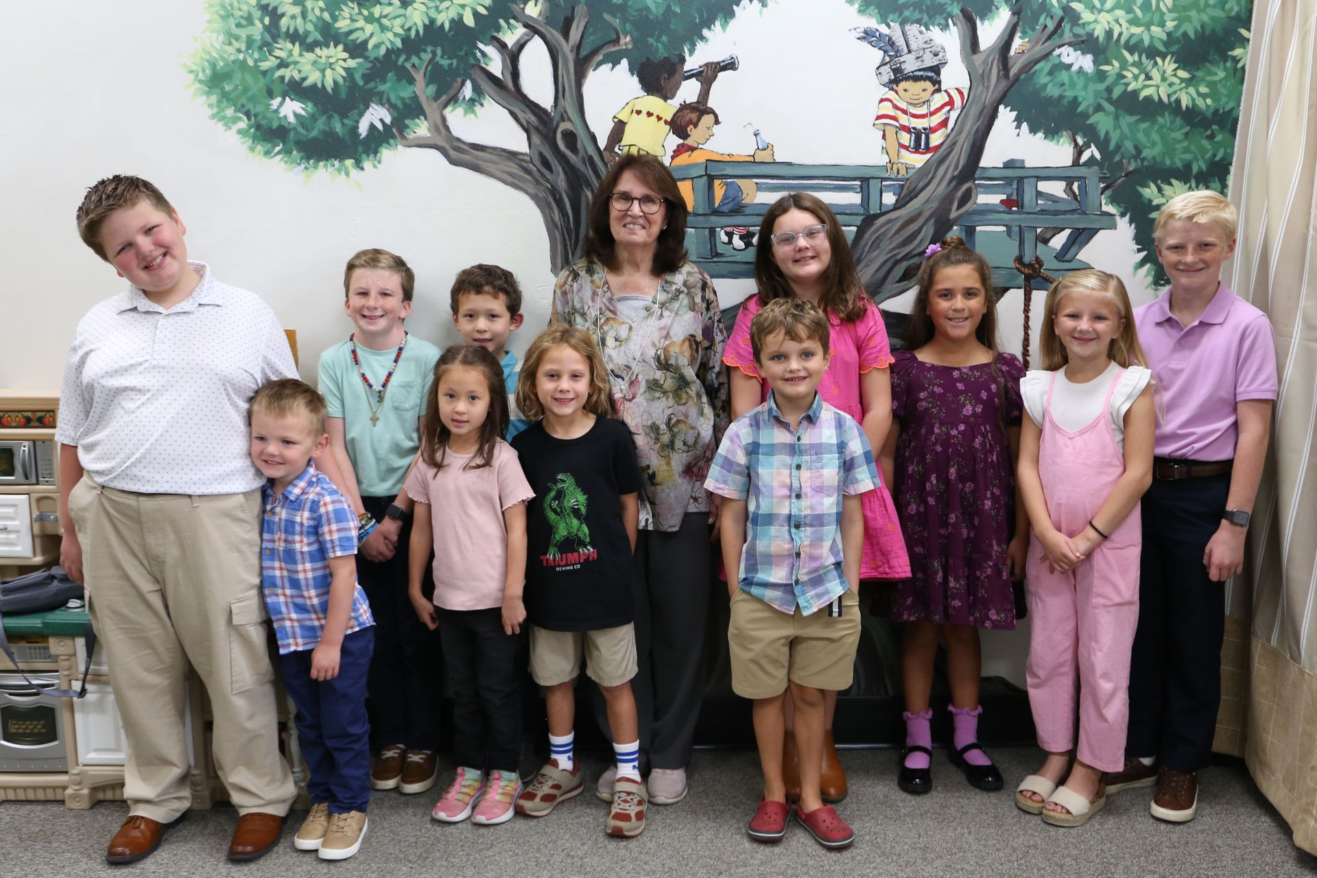 Group of children and two adults posing indoors in front of a painted tree mural.