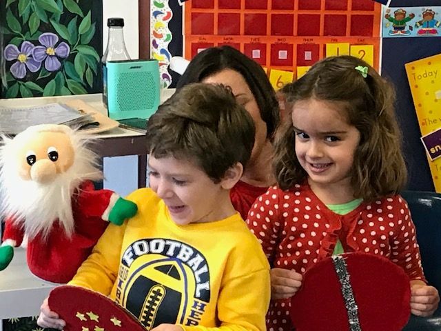 Two children in a classroom holding crafts beside a Santa puppet