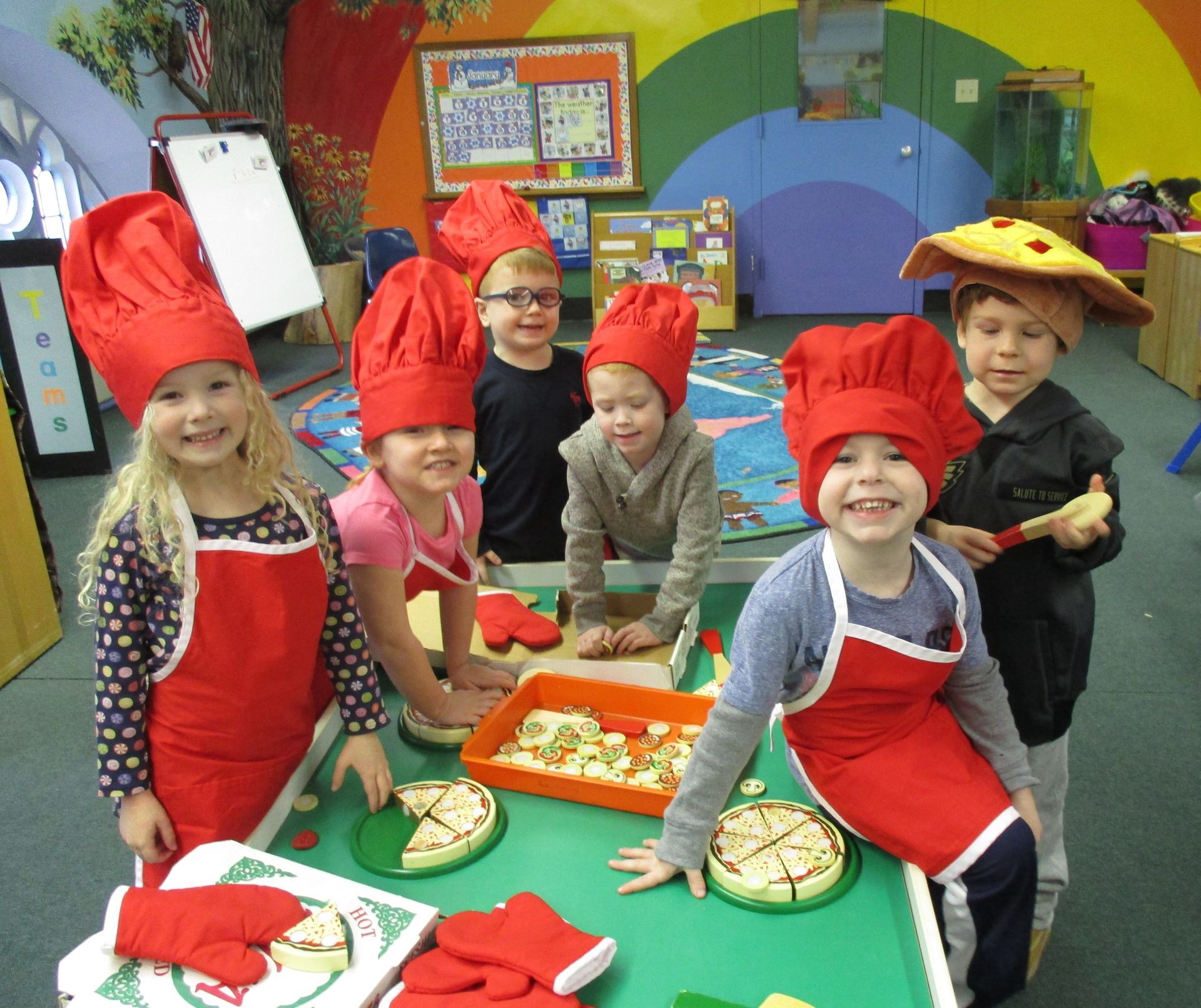 Children in red chef hats and aprons preparing food around a table in a colorful classroom