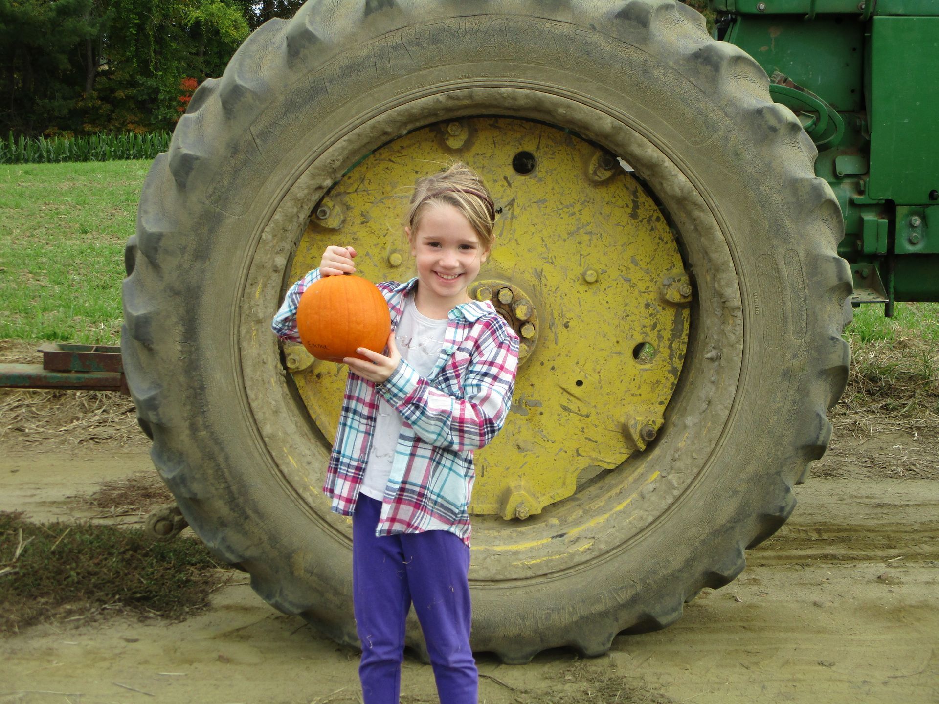 Girl holding an orange pumpkin in front of a giant tractor tire outdoors
