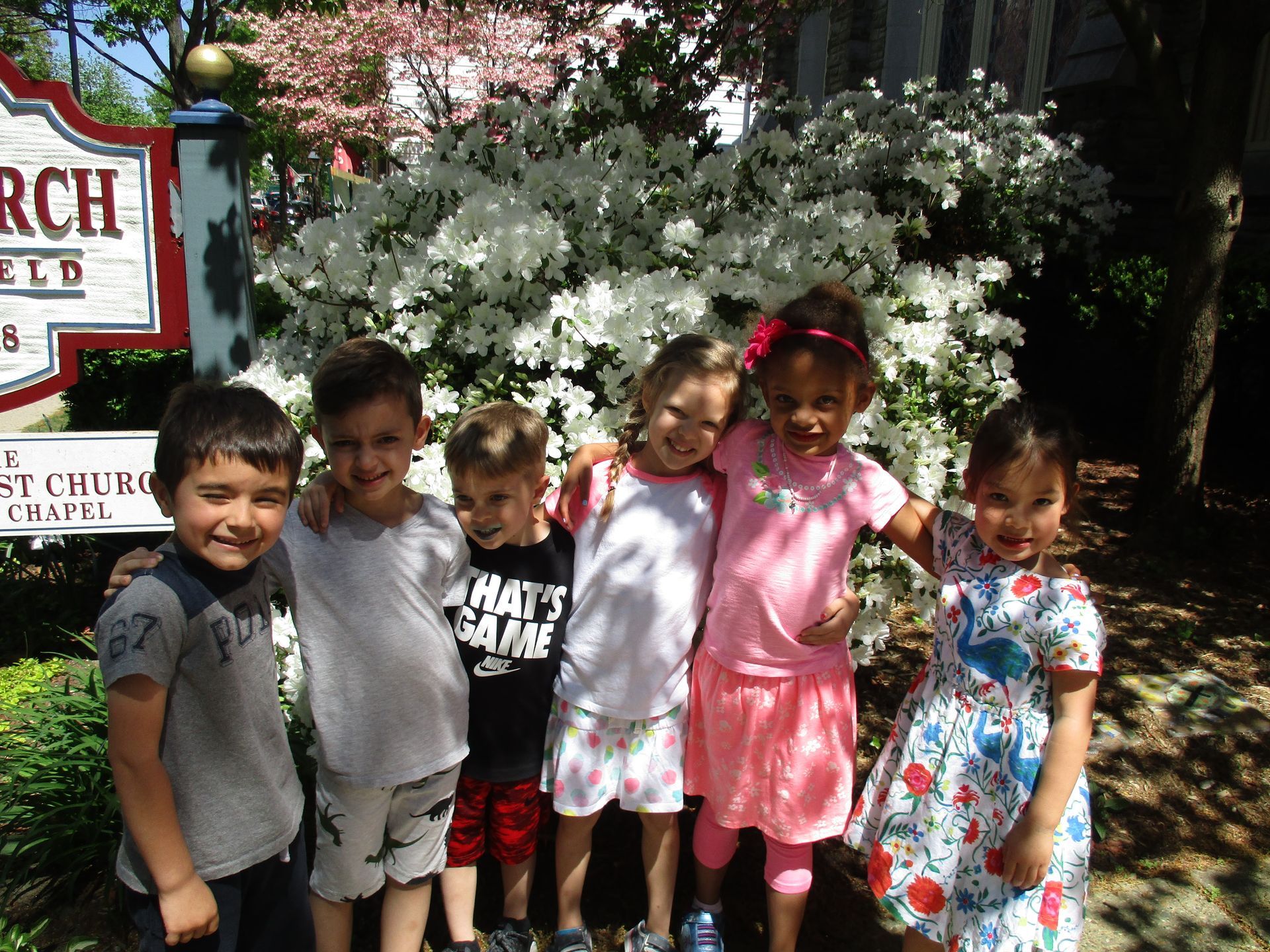 Five children smiling outdoors in front of flowering bushes and a church sign.