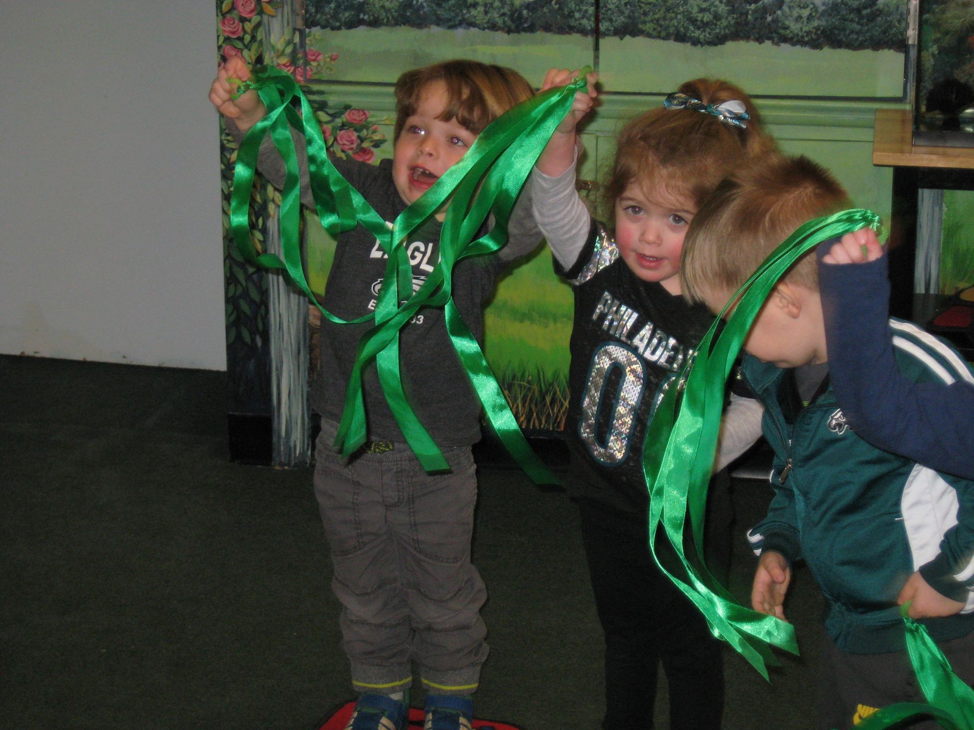 Two children holding green ribbons in a playroom, smiling and posing with raised arms