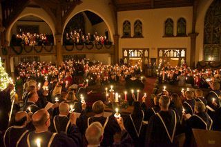 Crowded candlelit indoor gathering in a grand hall, with people holding candles and lights glowing throughout.