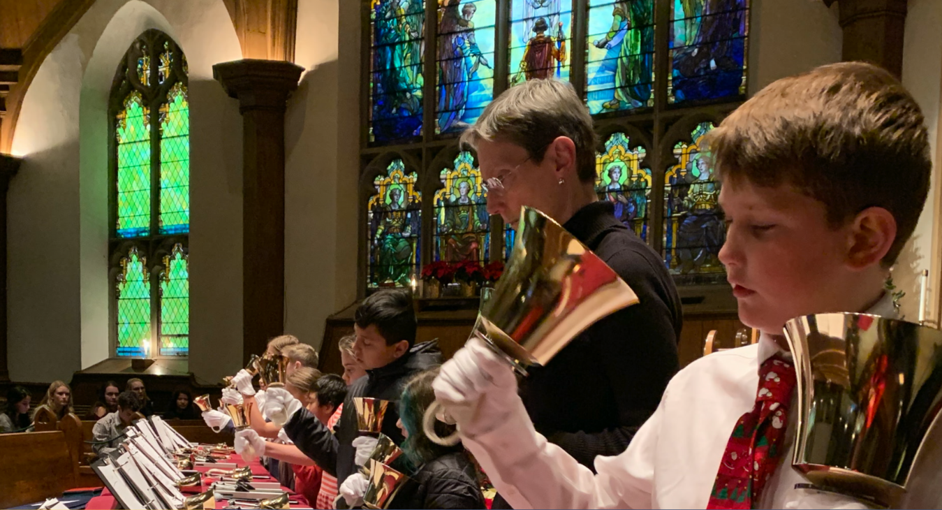 Children ring handbells during a church performance beneath stained-glass windows.