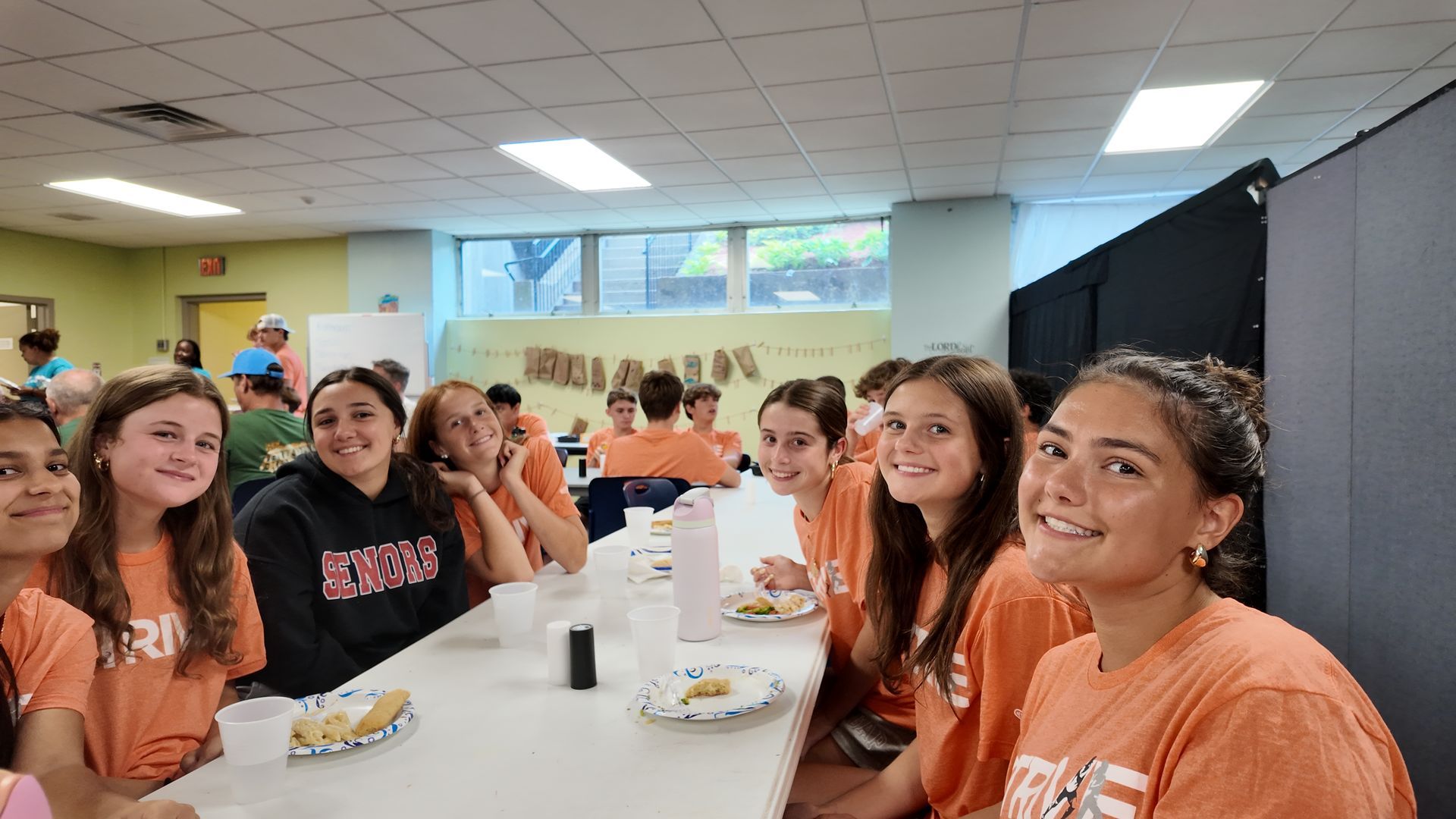 Group of students in orange shirts seated at a long table in a bright cafeteria, smiling at the camera.