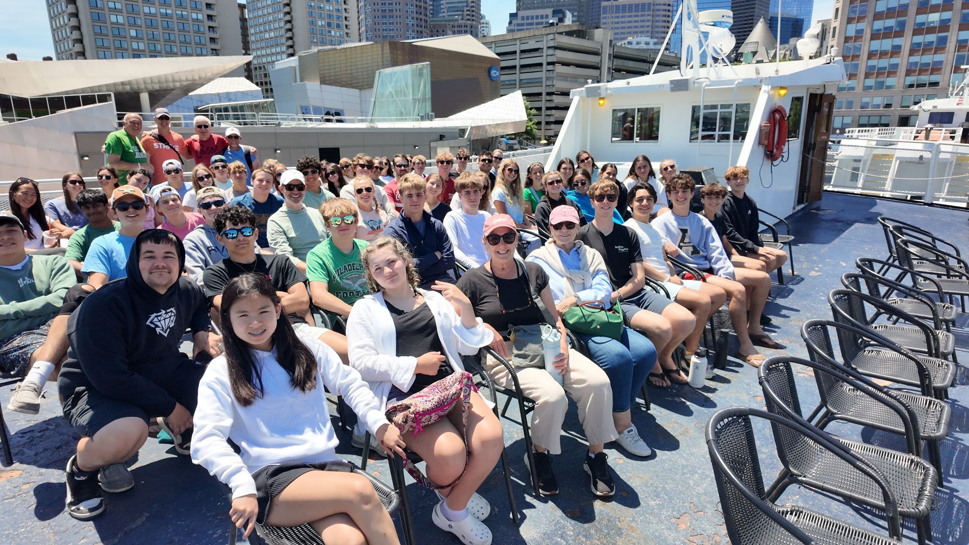 Crowded boat deck with passengers seated in rows, smiling in bright sunlight near city buildings.