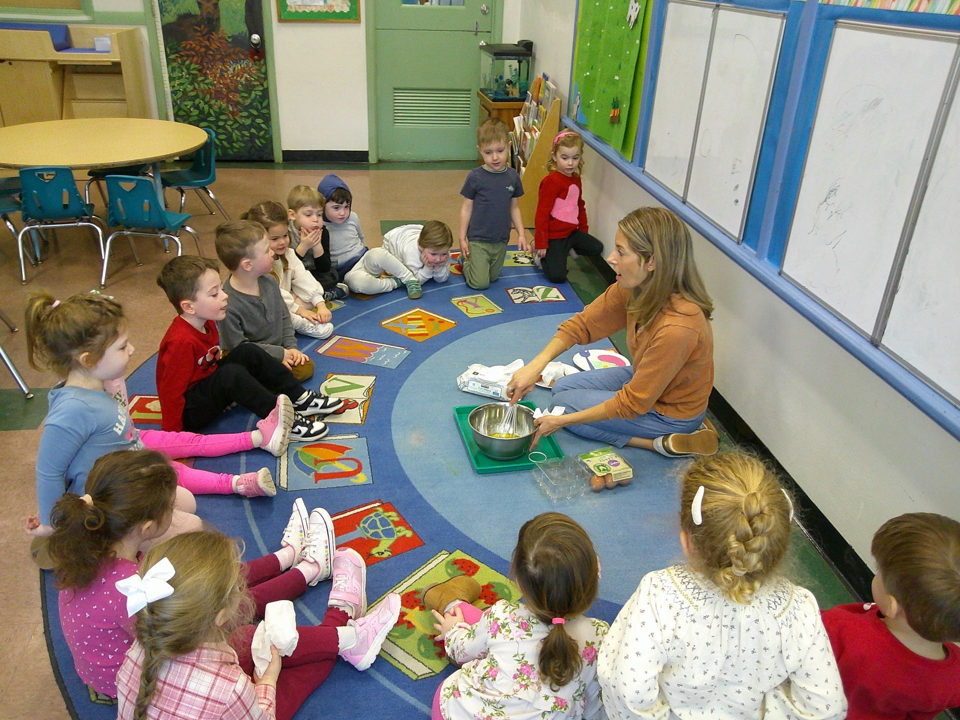 Children sit in a circle on a classroom rug while a teacher leads an activity with a bowl in the center.