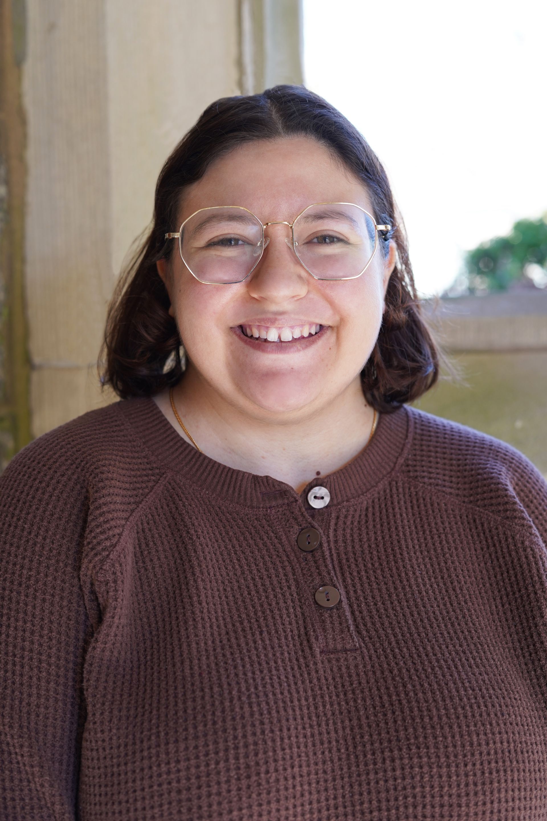 Smiling person in glasses and a brown sweater standing outdoors.