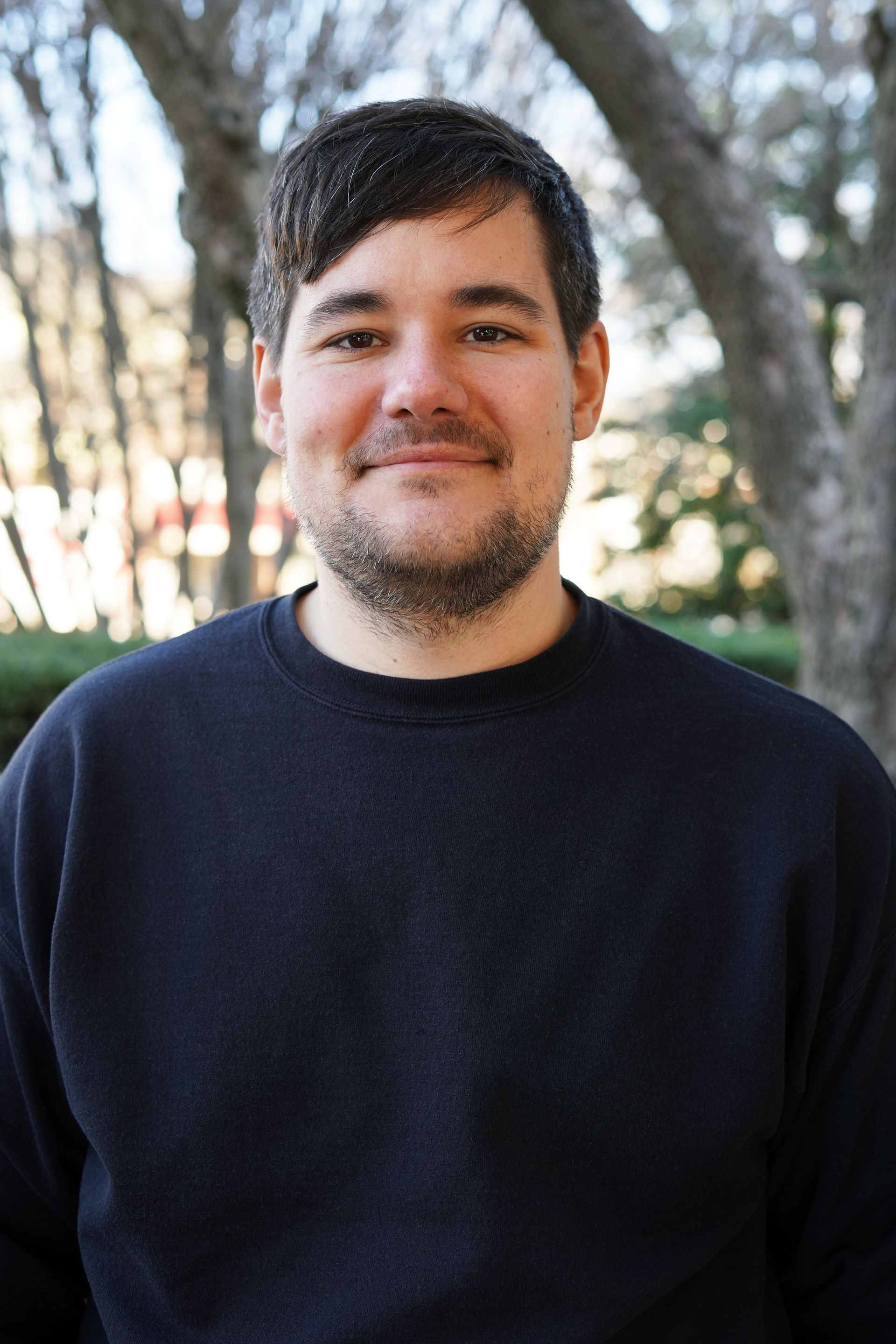 Man standing outdoors in a dark sweater, smiling softly with trees blurred in the background