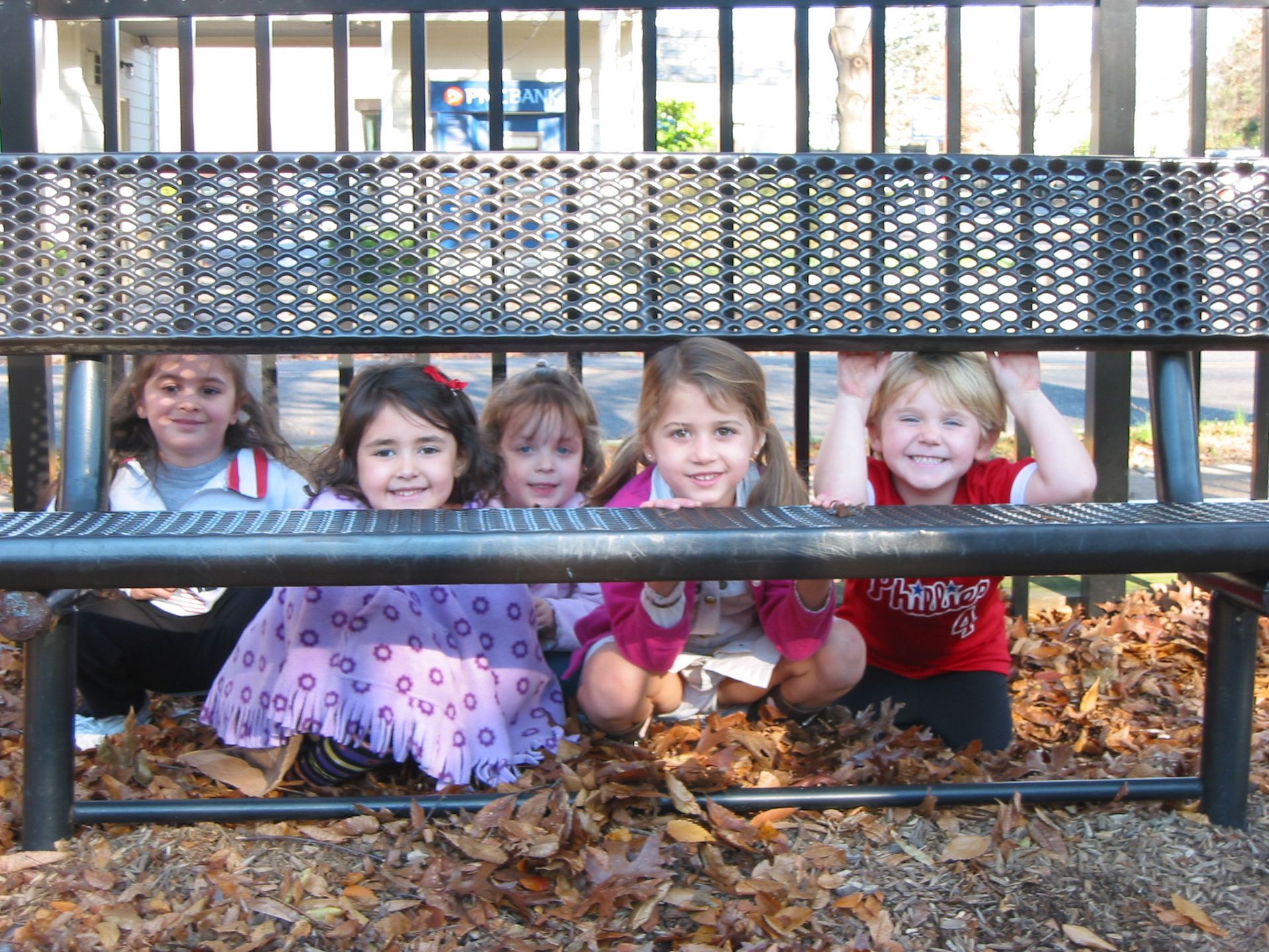 Five children peek through a metal fence, smiling in a playground with autumn leaves.