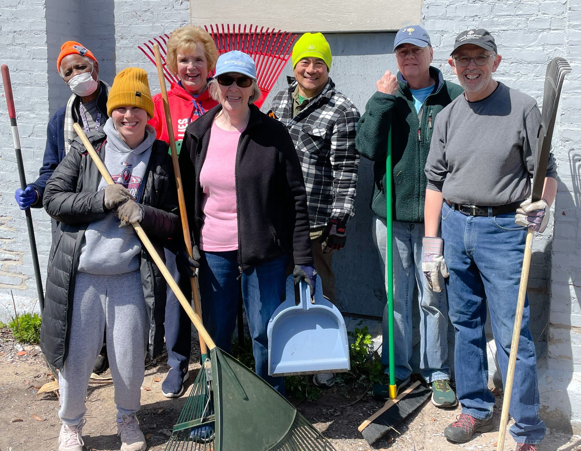 Group of seven volunteers with rakes and shovels standing in front of a wall outdoors.