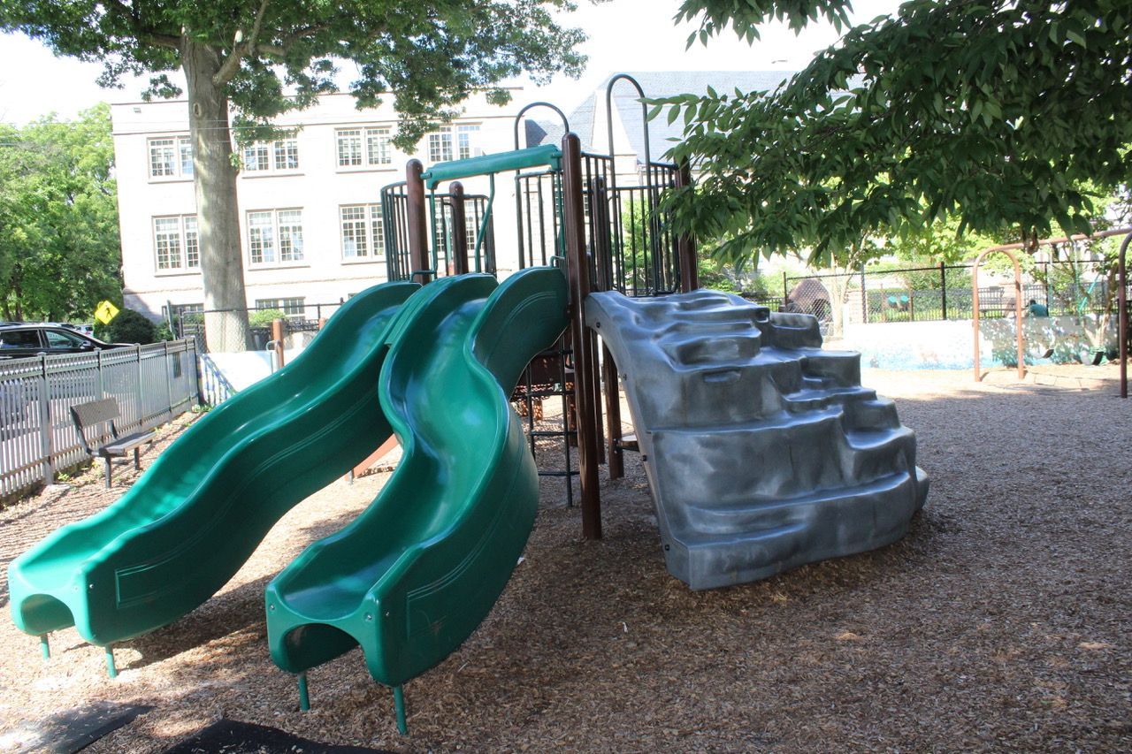 Two green slides and a gray climbing wall in a playground under trees