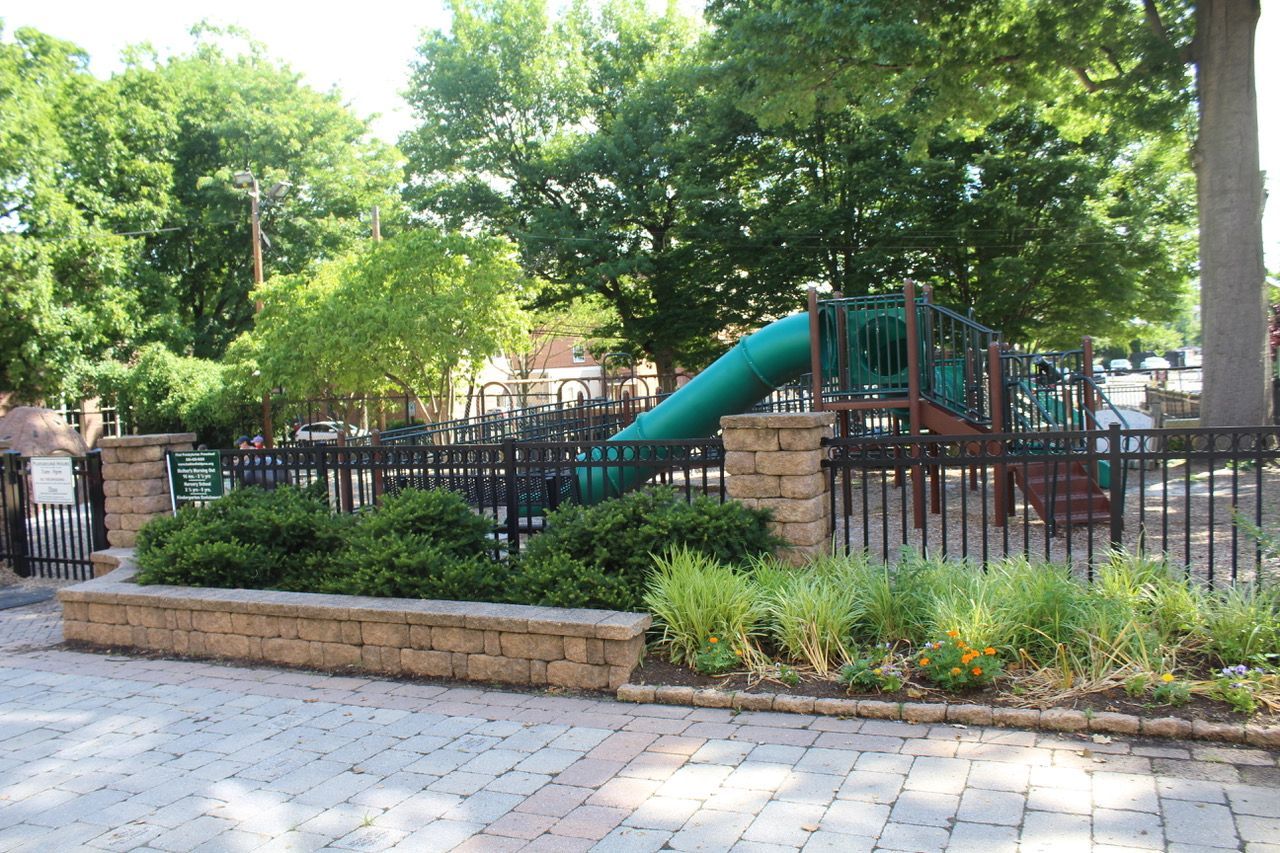 Tree-lined playground with a green slide, black fence, and stone edging in a park