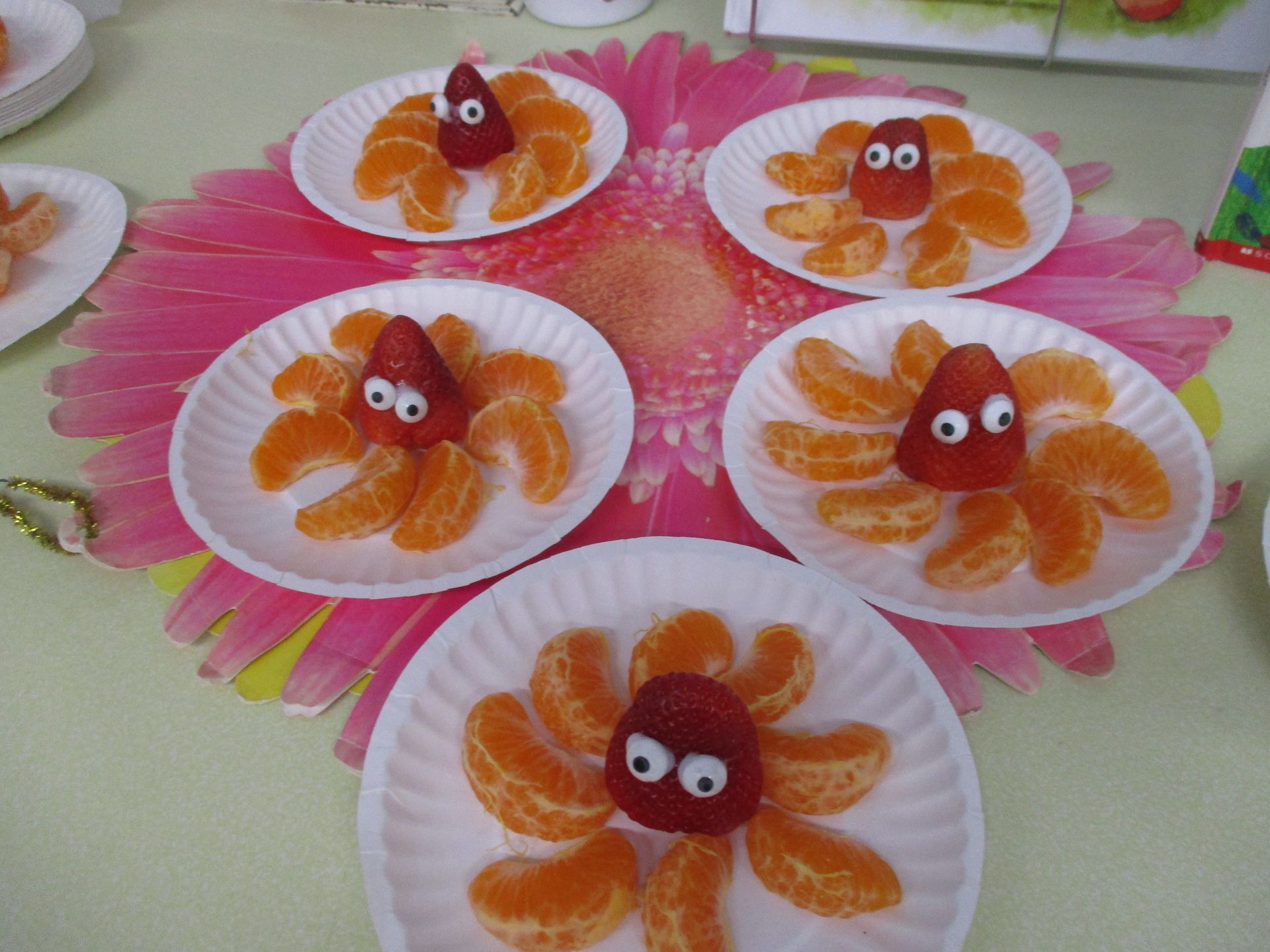 Five plates of orange-slice octopus snacks with googly eyes on pink flower mats
