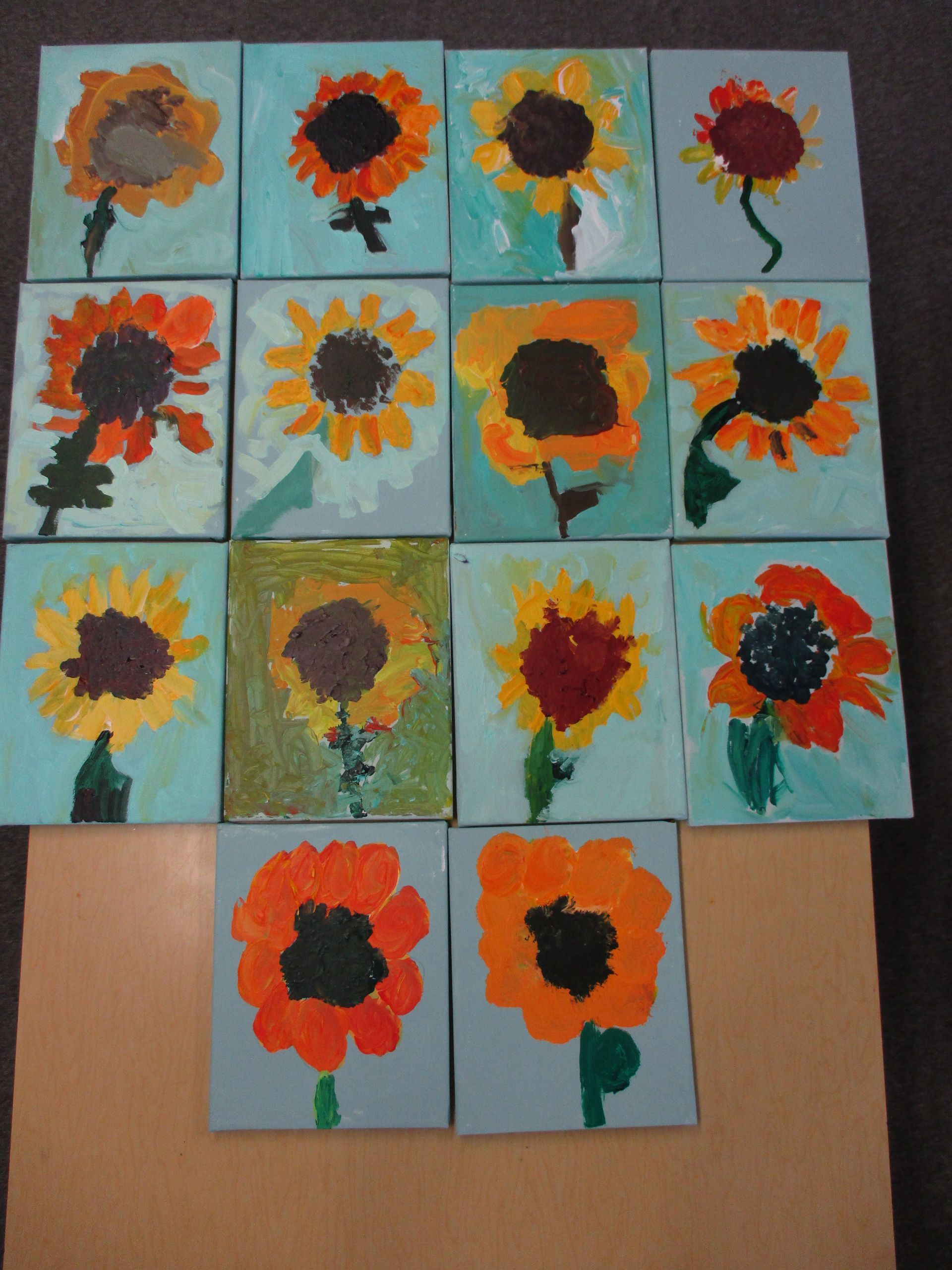 Handmade sunflower paintings arranged in a grid on blue paper tiles against a tan background