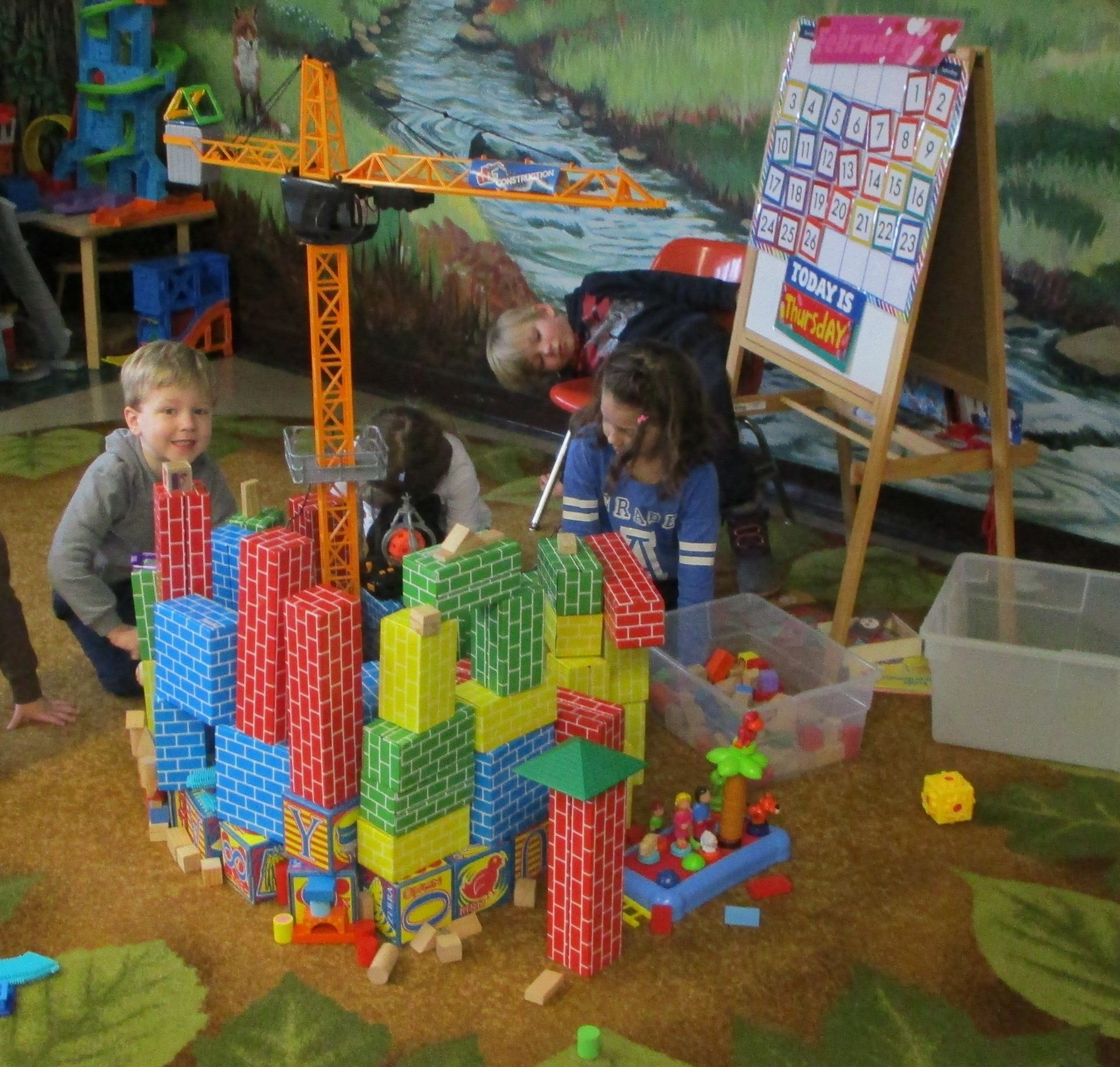 Children building colorful block structures in a playroom with a crane toy and easel.