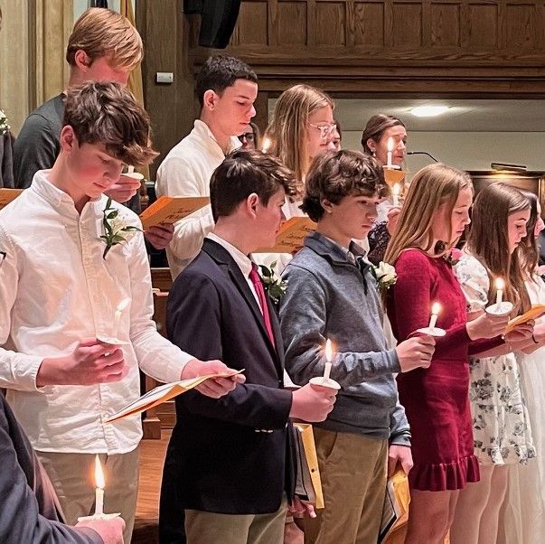 People holding lit candles in a ceremony inside a church or hall, with one in a suit and others in dress clothes
