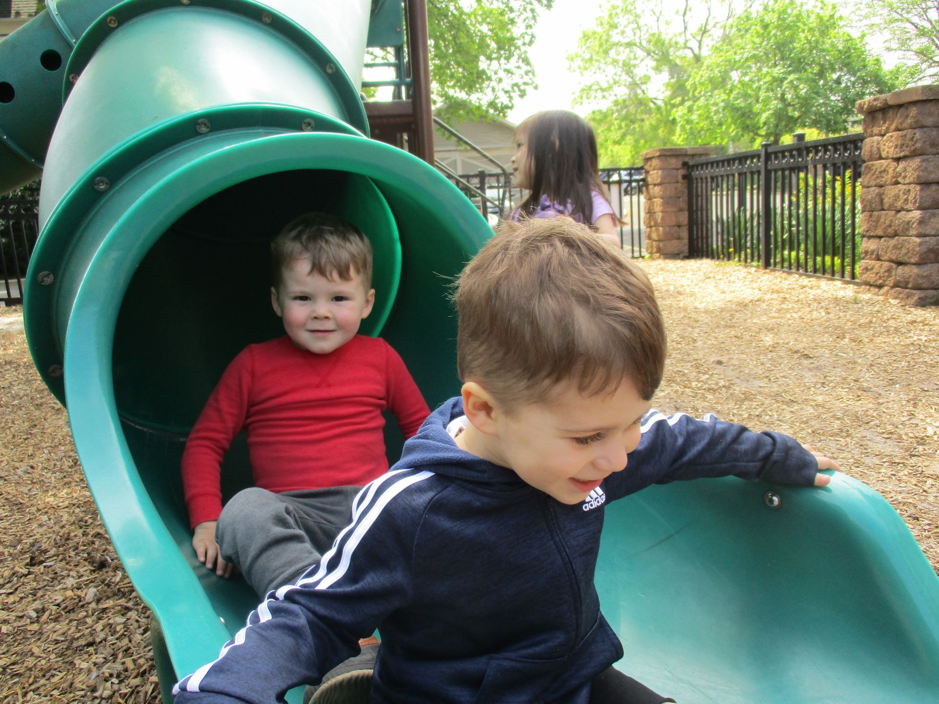 Two children on a green playground slide, one sitting inside and one climbing out at the bottom.