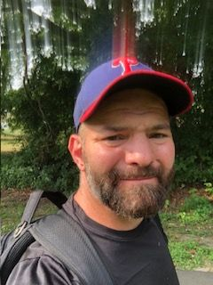 Smiling man in a navy and red cap taking a selfie outdoors near trees and grass