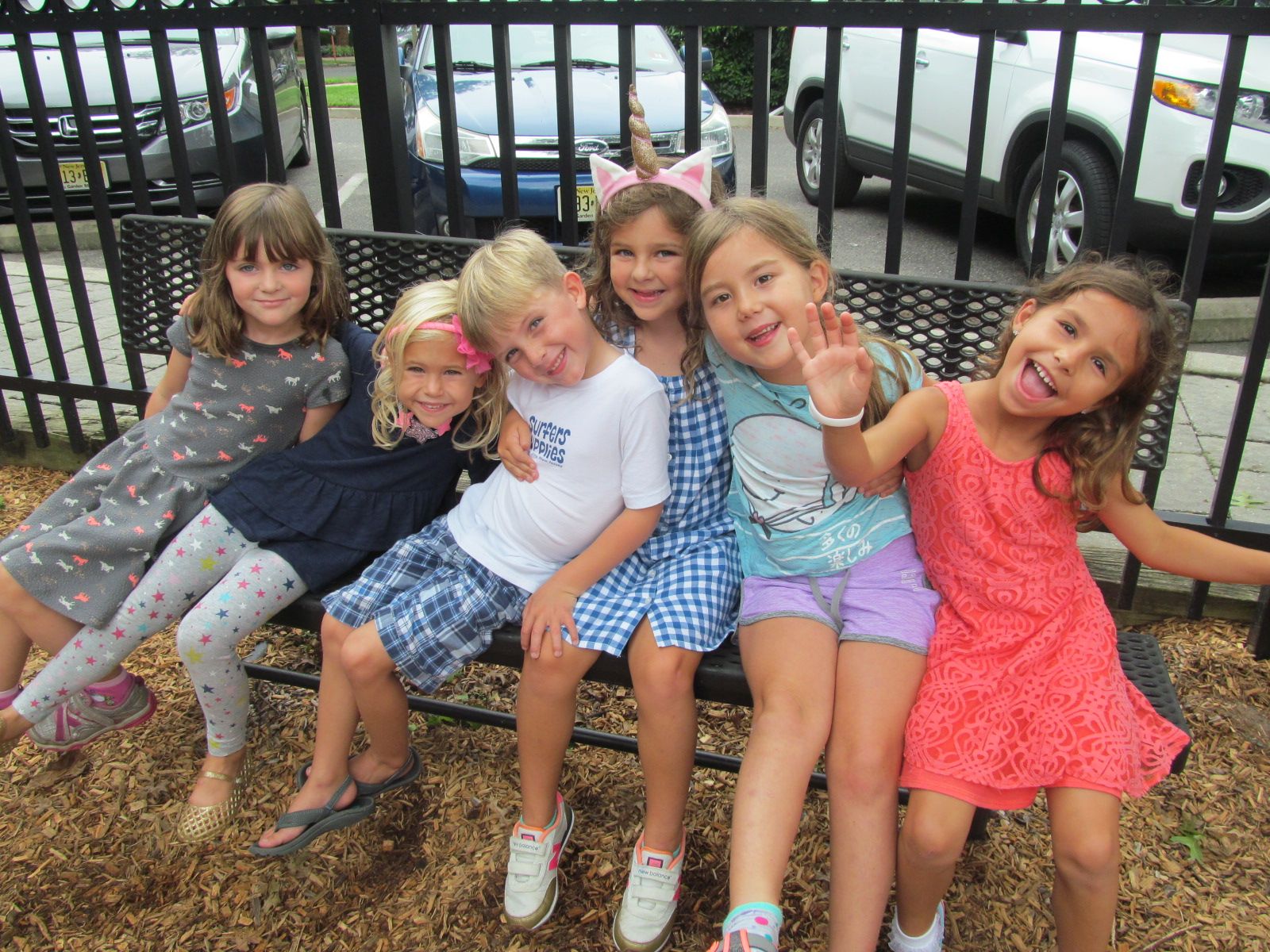 Five children sitting on a bench, smiling and waving outdoors near a fence.