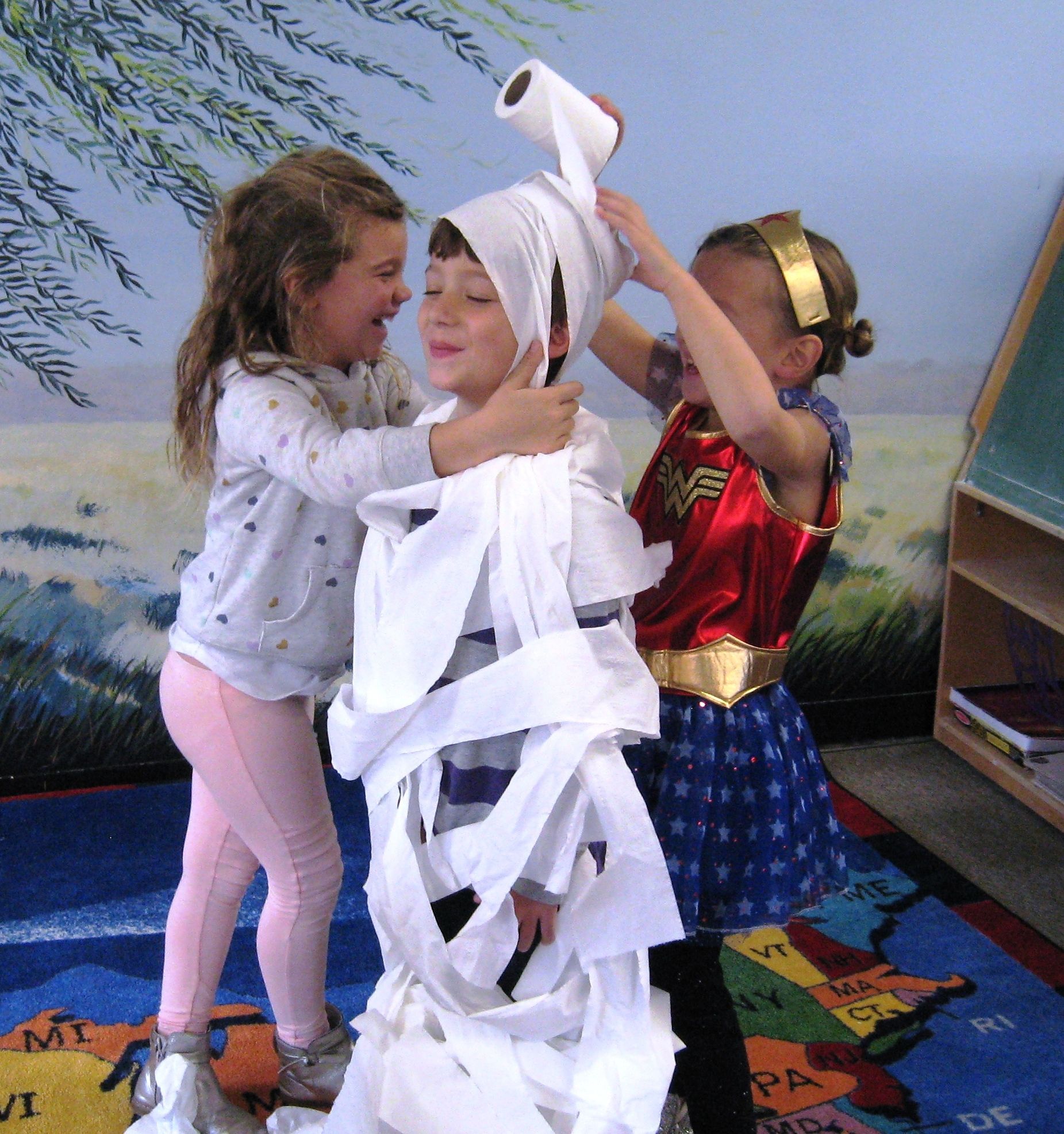 Three children play dress-up indoors, wrapping one child in white toilet paper mummy costume.