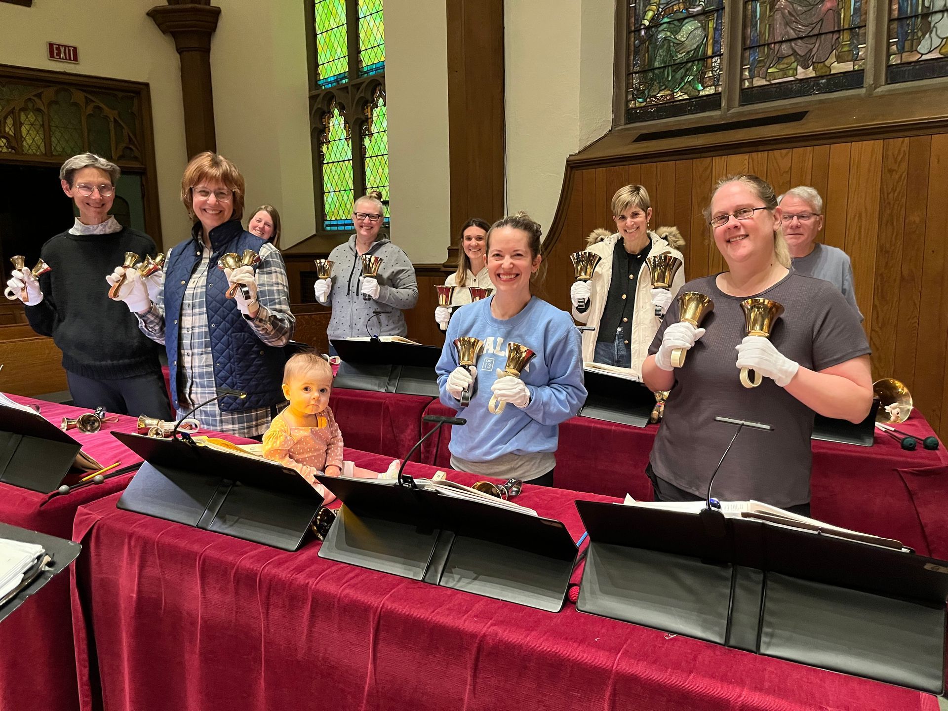 People in a church hold handbells behind red-draped tables, smiling during a rehearsal.