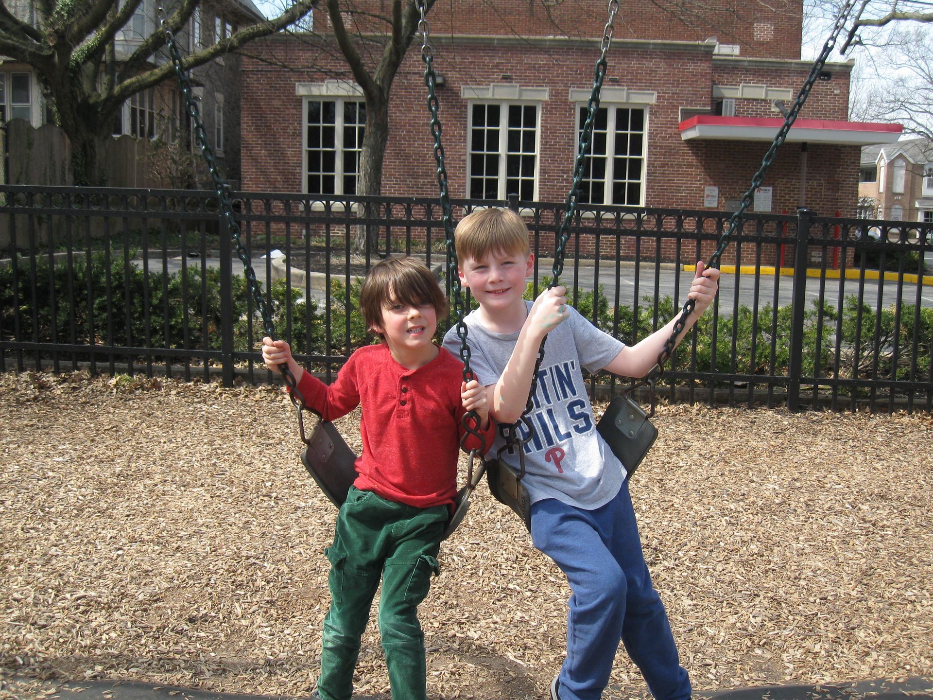Two children on swings in a playground, smiling in front of a brick building.