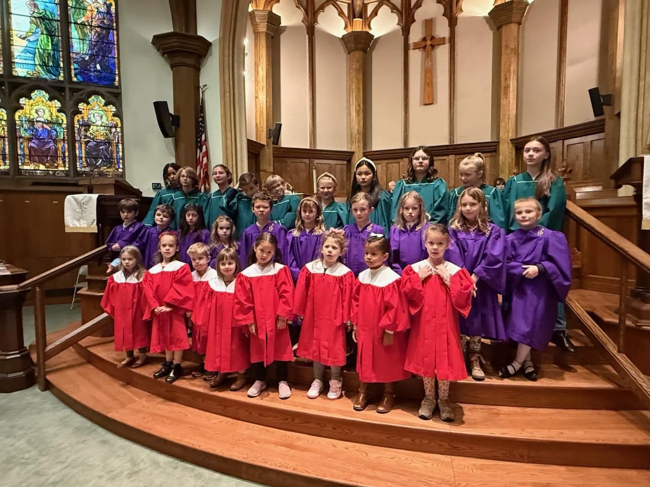 Choir children in red and purple robes standing on church steps under stained glass windows