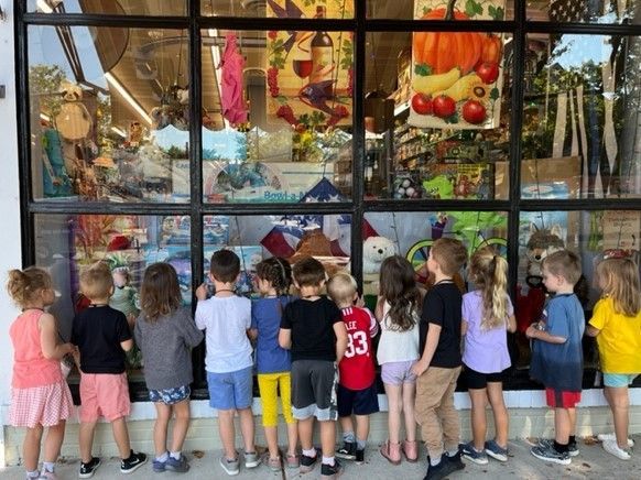 Children standing outside a shop window, looking at colorful fruits and hanging decorations inside.