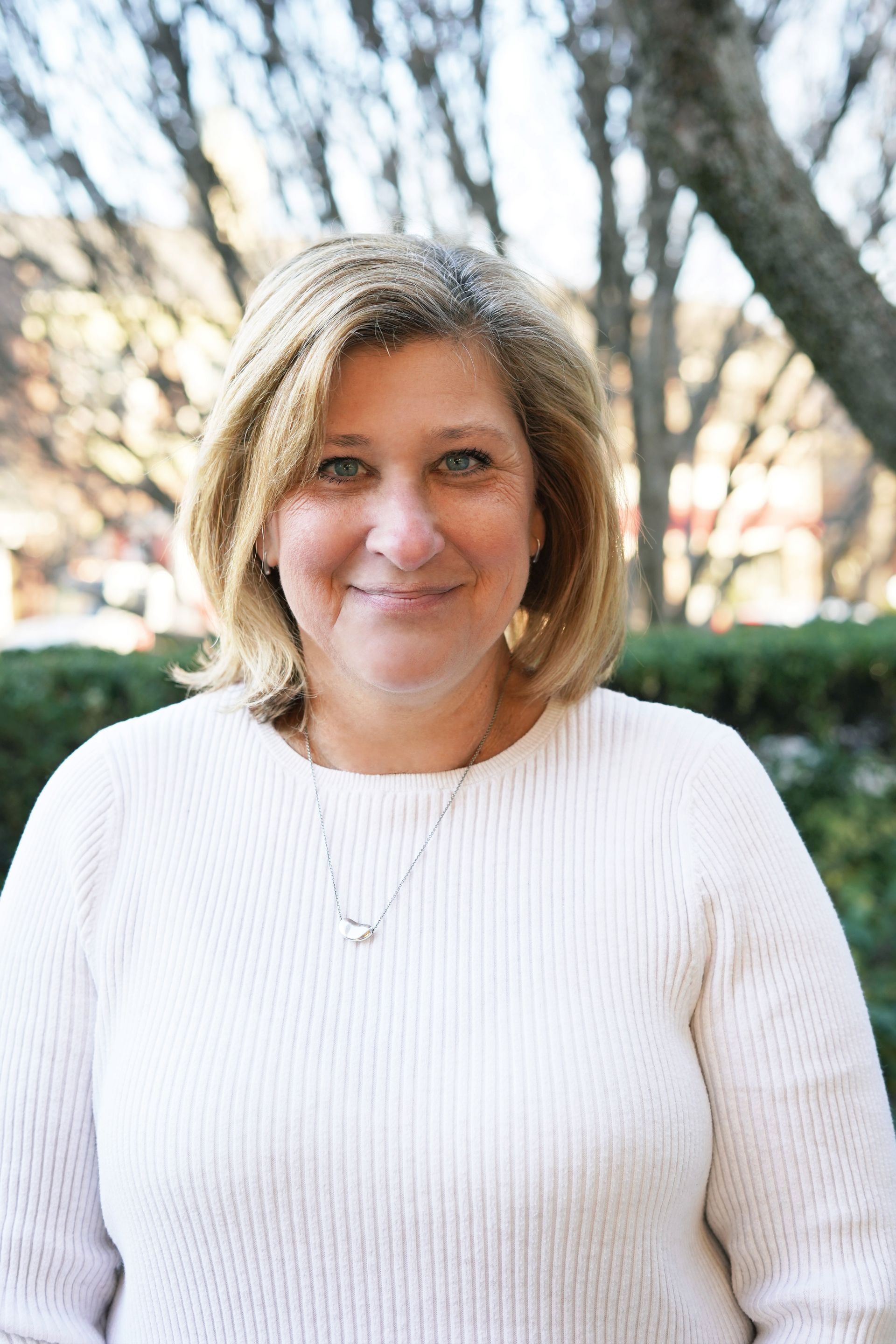 Person in a white sweater smiling outdoors with trees in the background