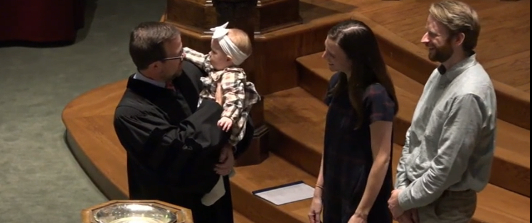 Four people stand in a wood-paneled hall while one holds up a baby wrapped in white.