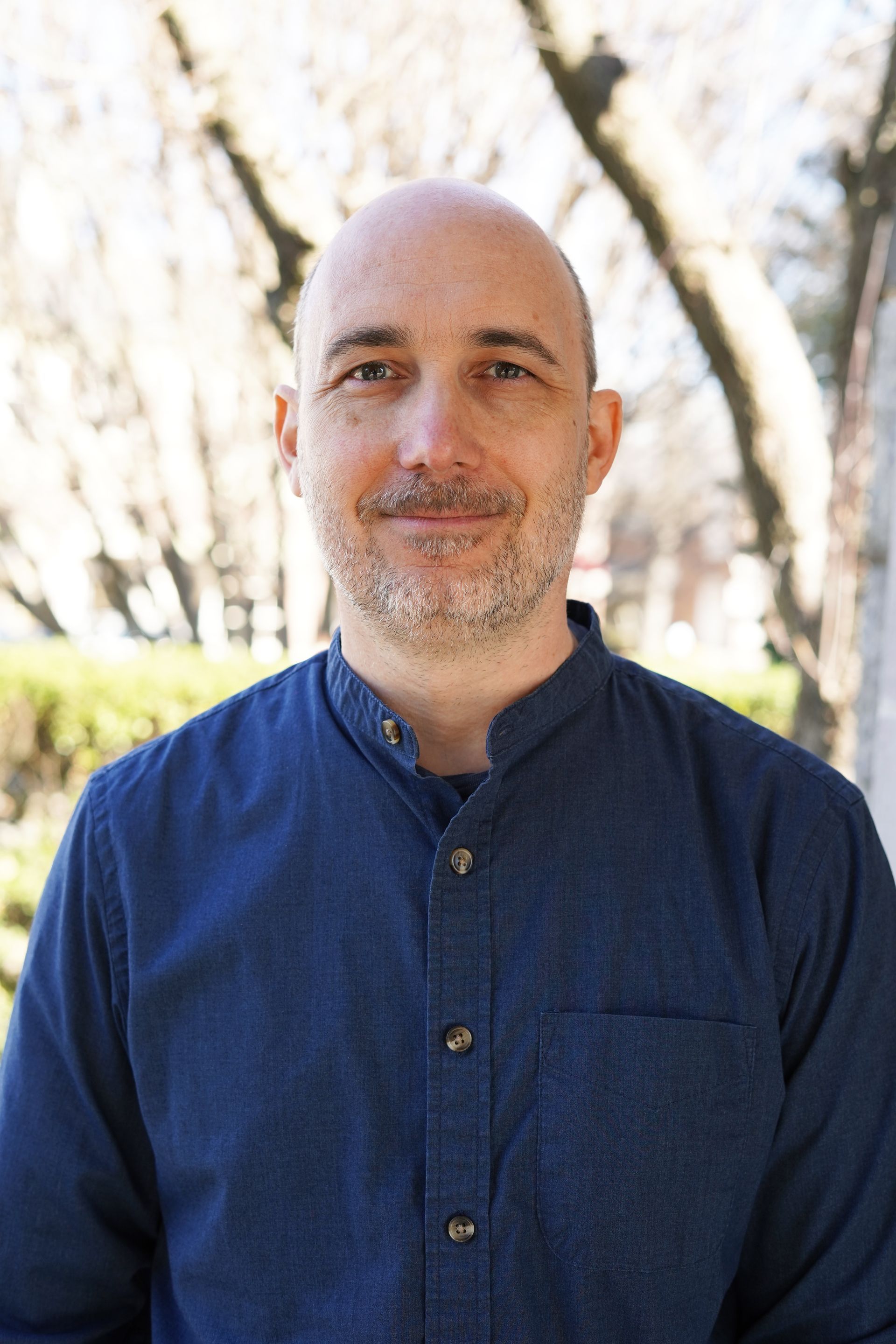 Smiling bald man in a blue shirt standing outdoors with blurred trees in the background
