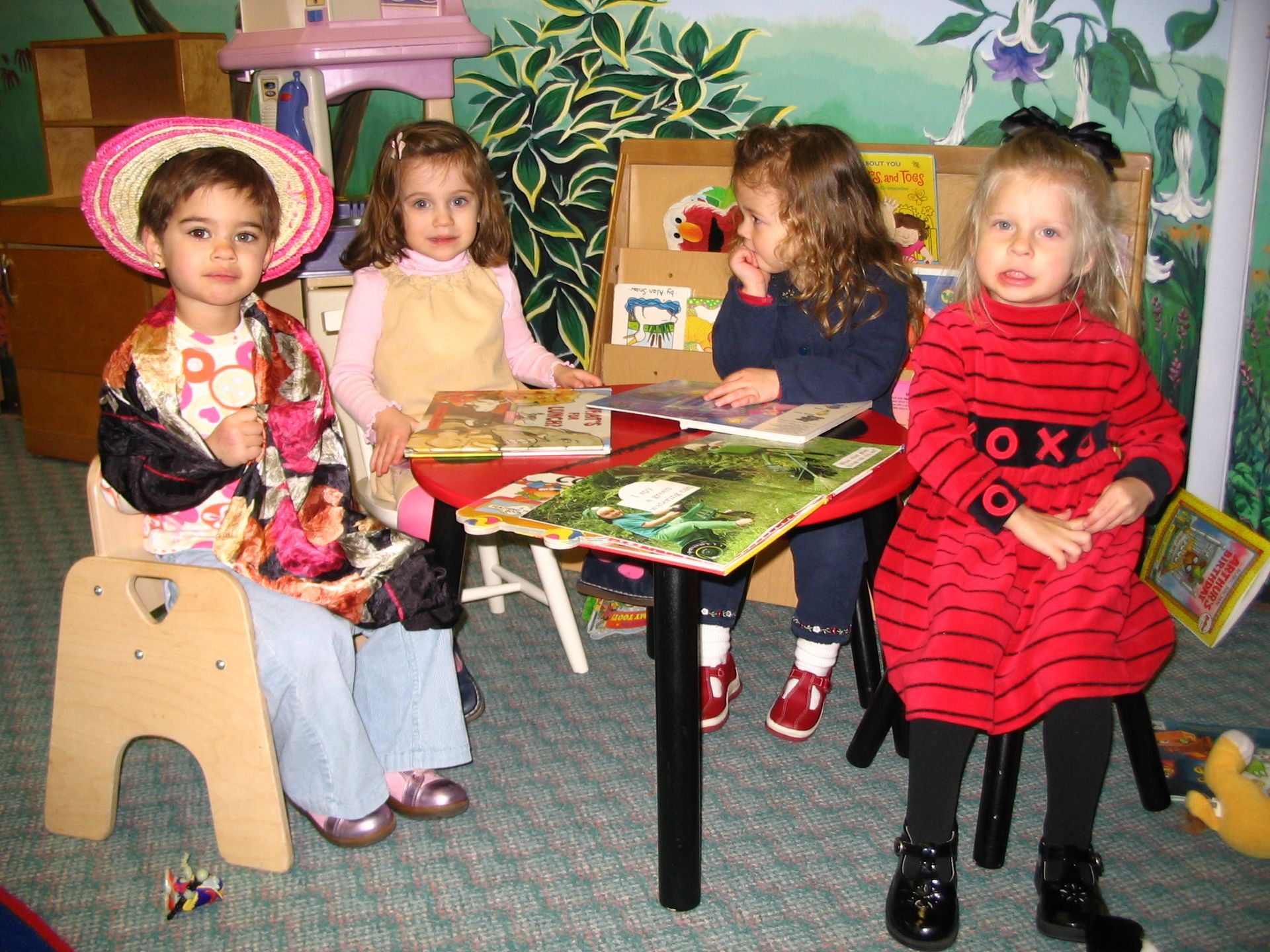 Four children sit around a small table in a colorful playroom, surrounded by toys and painted murals.