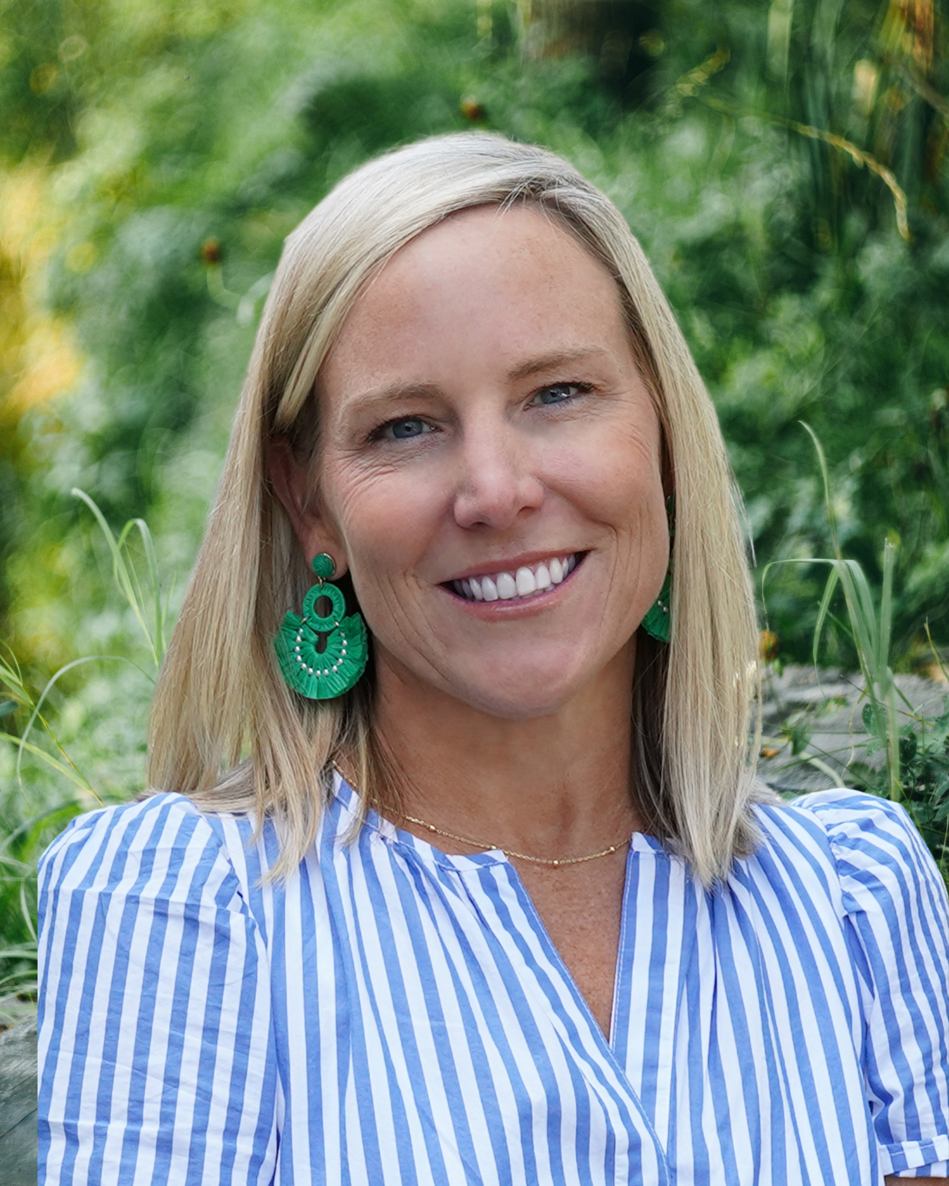 Smiling woman in a blue-and-white striped blouse with green earrings, standing outdoors against greenery
