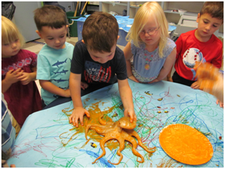 Children gathered around a blue table touching an octopus and sea-life drawings in a classroom