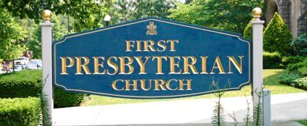 First Presbyterian Church sign in gold letters on a blue background, set by a walkway with greenery