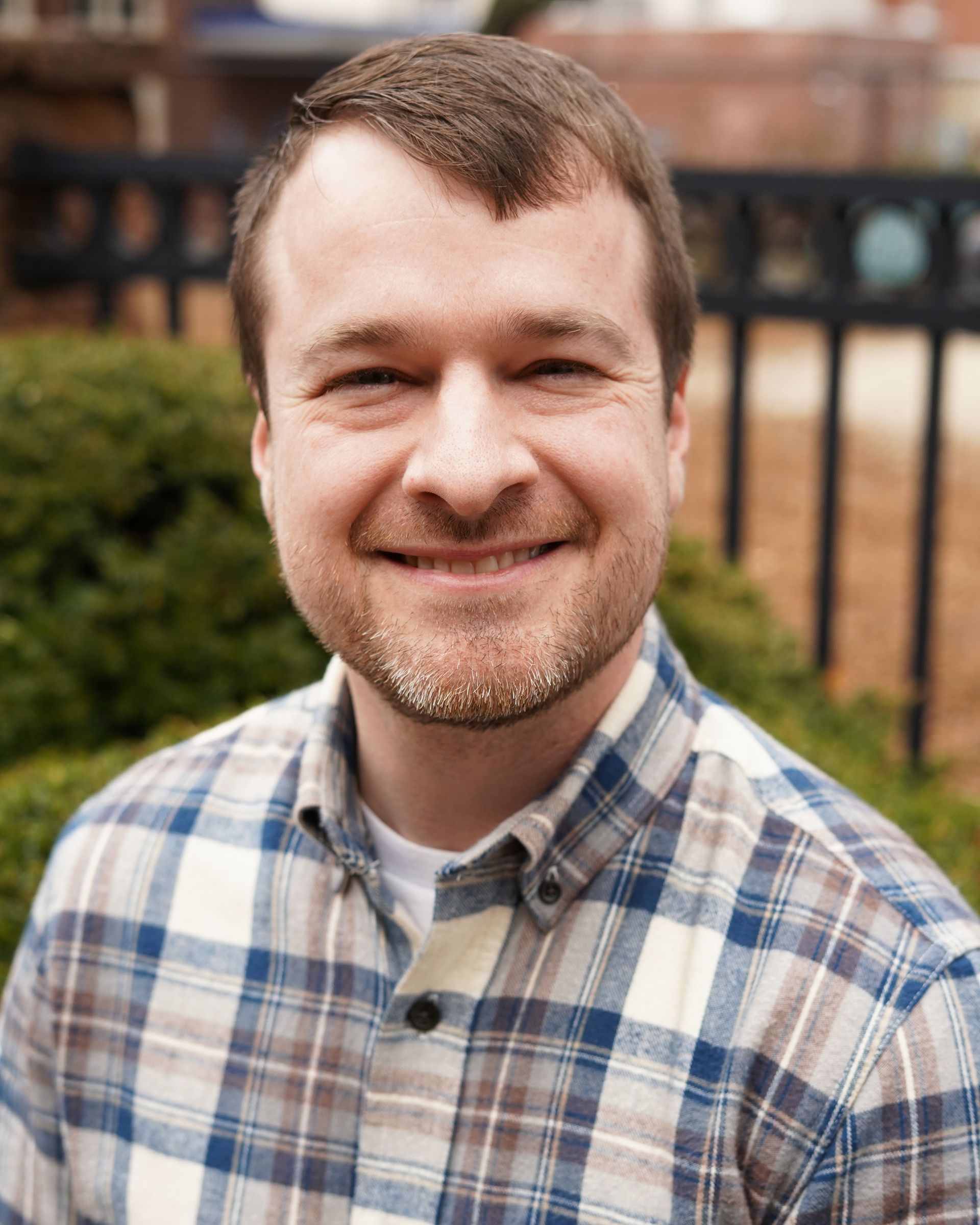 Smiling man in a plaid shirt standing outdoors in front of a building and fence
