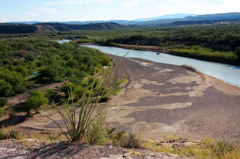 River winding through a dry, rocky valley with green shrubs and distant mountains under a clear sky