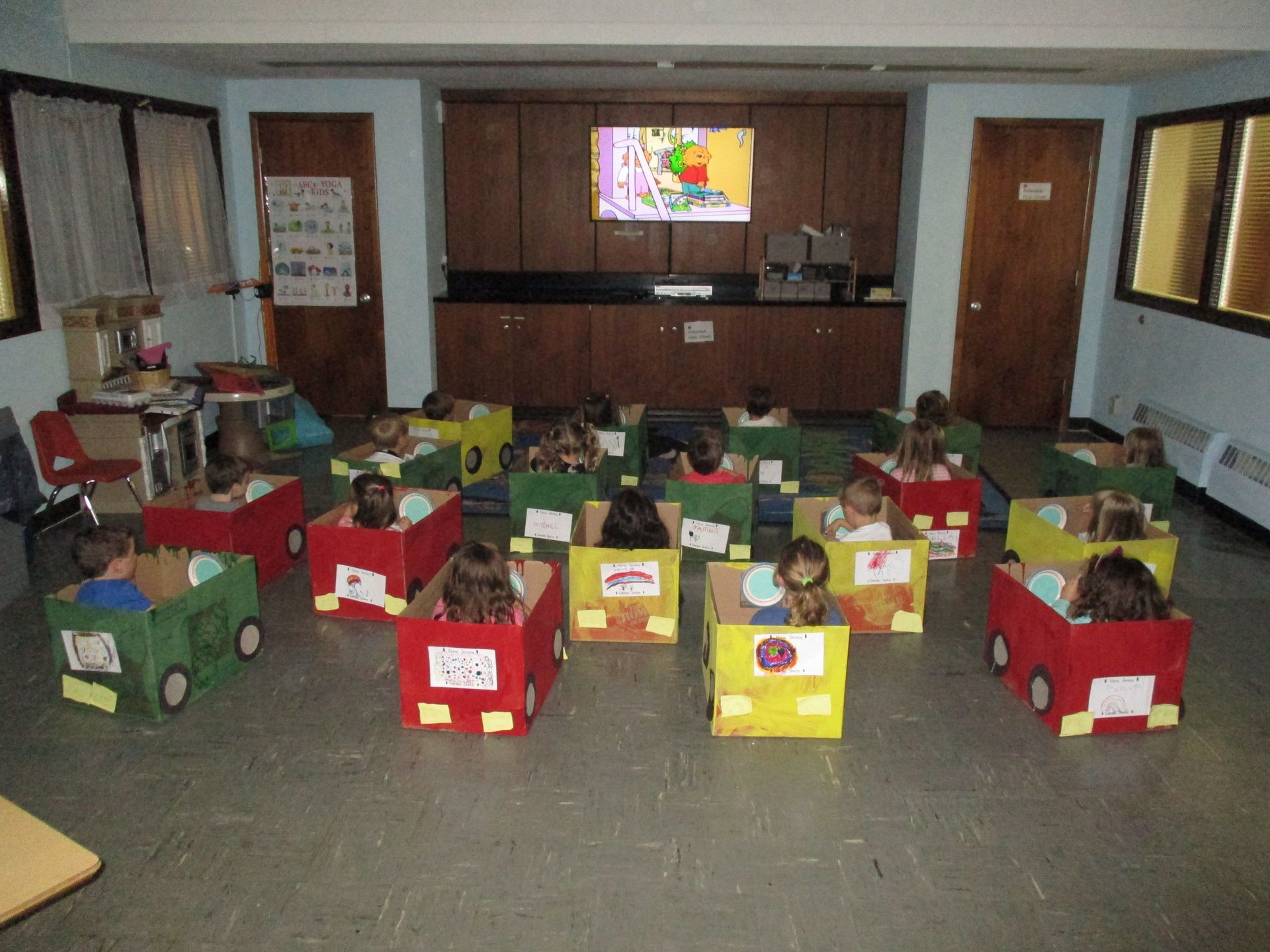 Children in boxy race car costumes sit in a classroom facing a wall-mounted screen.